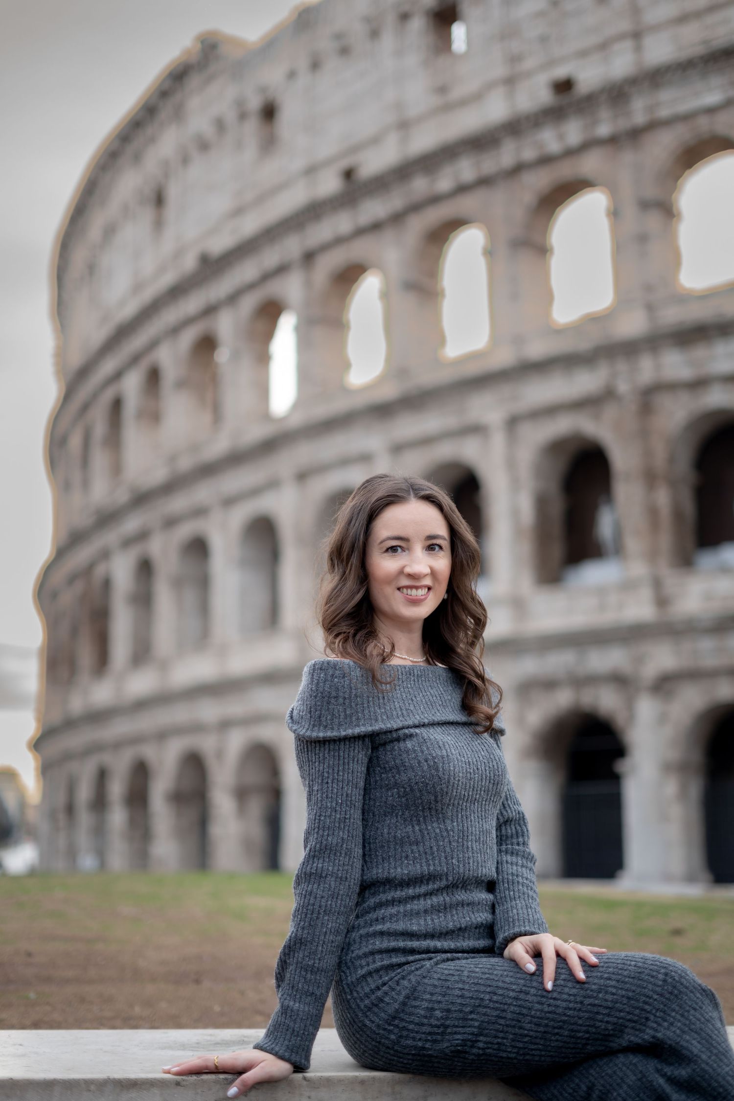Young woman in gray knit dress sitting in front of the Colosseum in Rome, Italy, smiling at the camera.