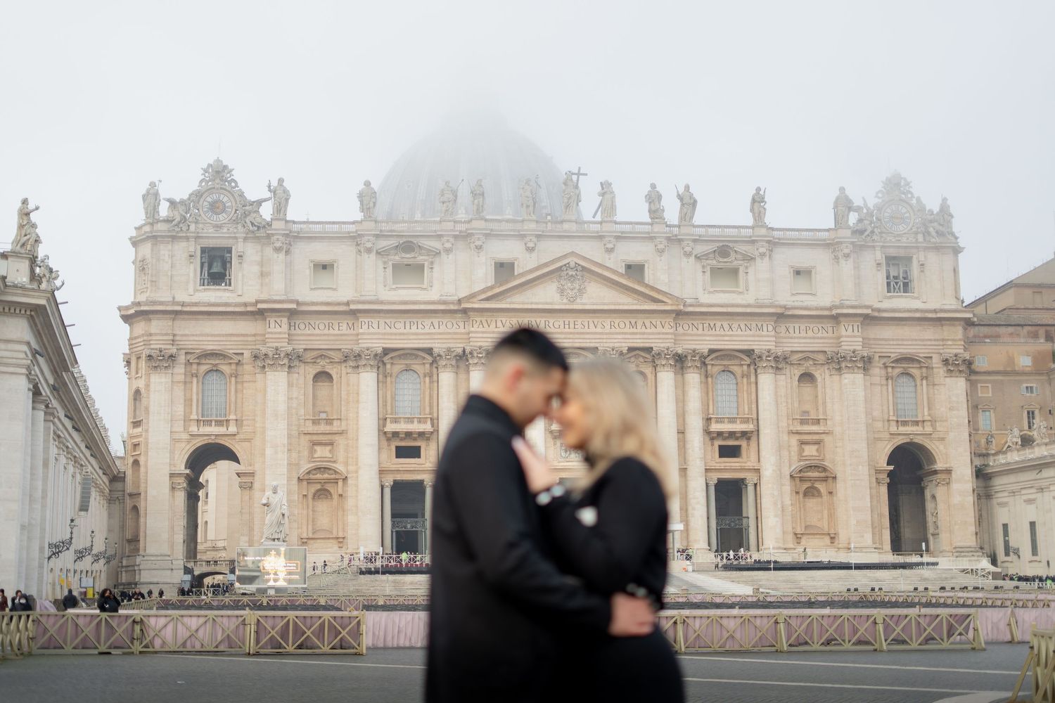 Couple embracing in front of St. Peter's Basilica in Vatican City on a misty day.