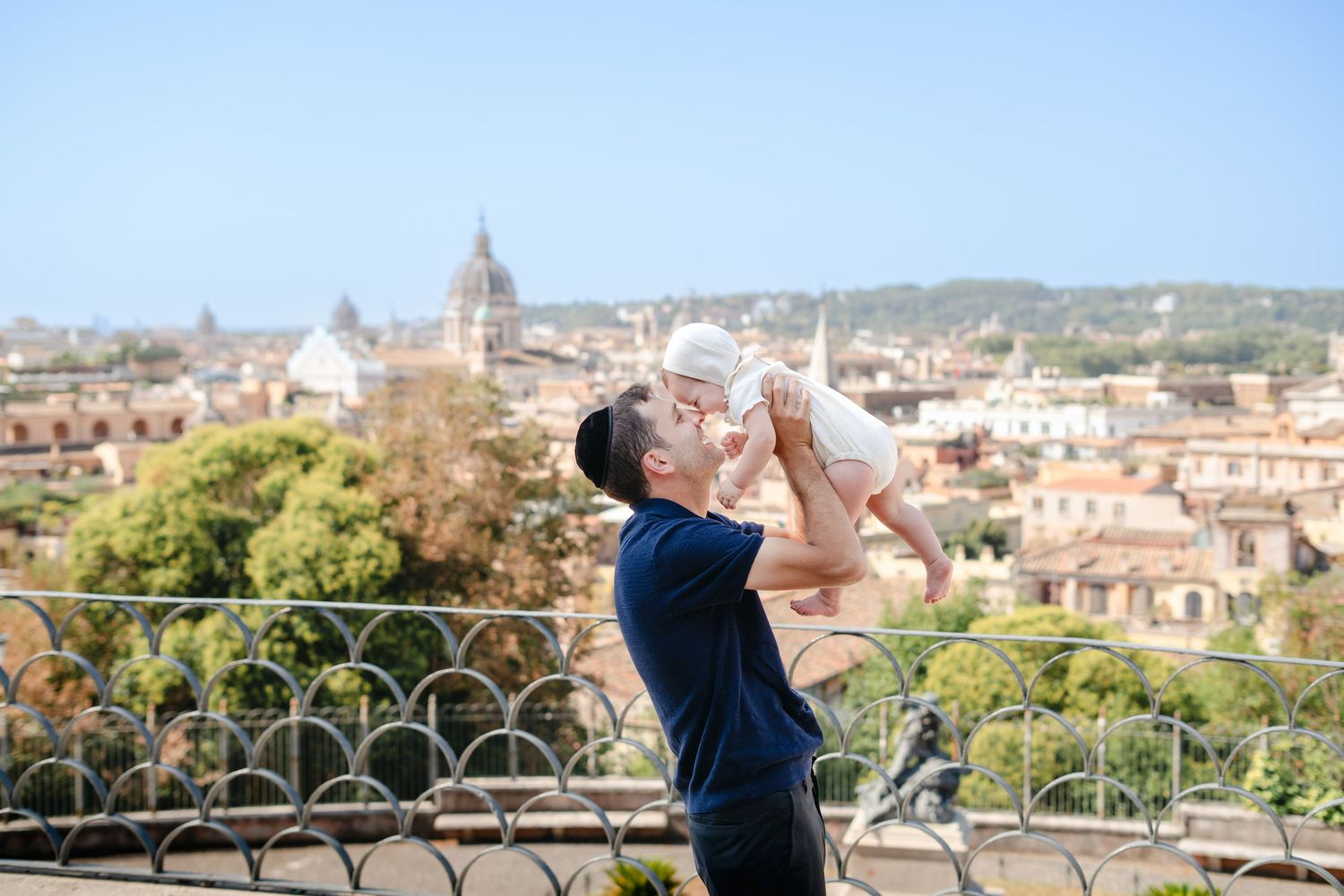 Father lifting baby in the air with Rome cityscape and domes in the background on a sunny day.