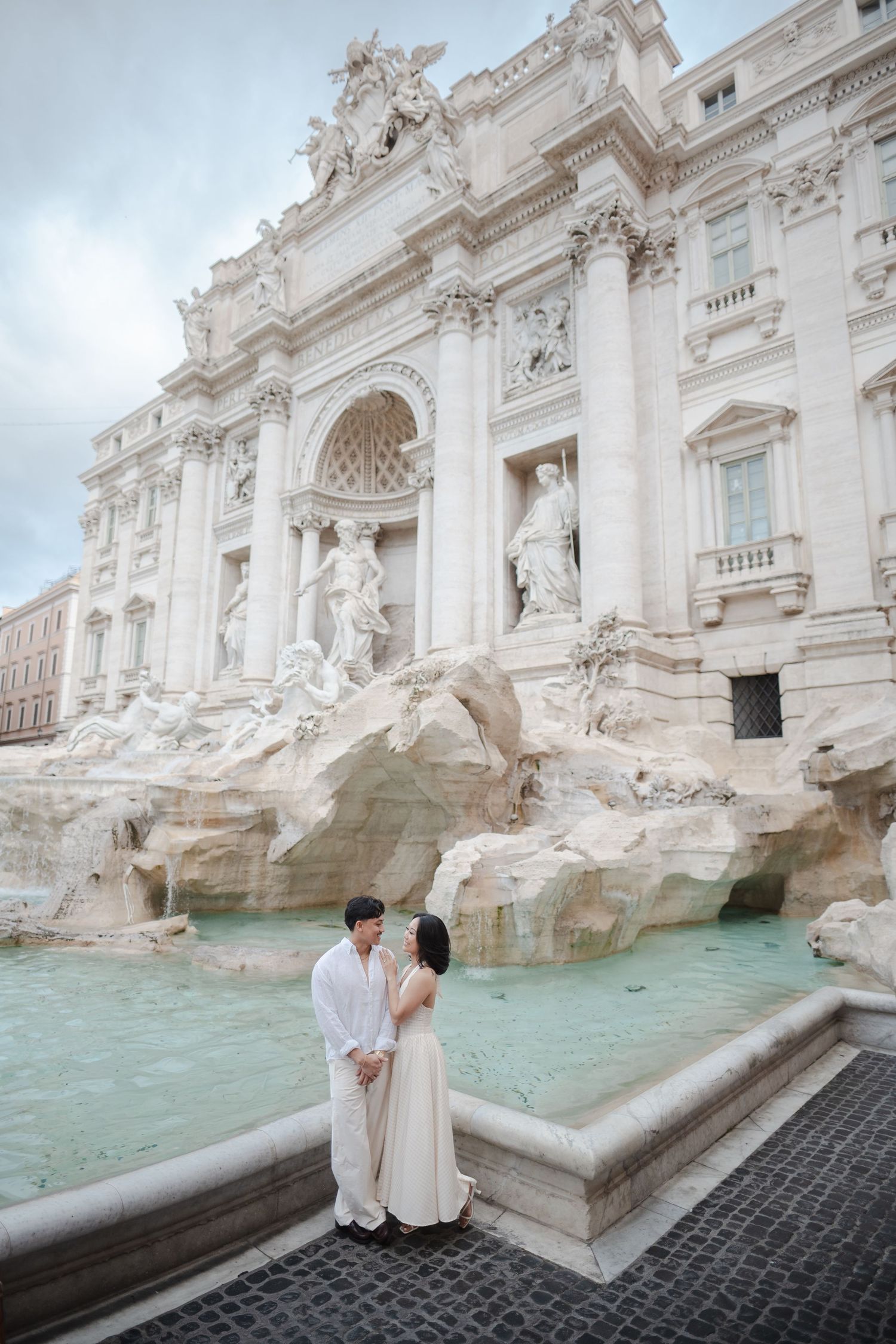 Couple in elegant attire standing romantically before the Trevi Fountain in Rome, Italy.