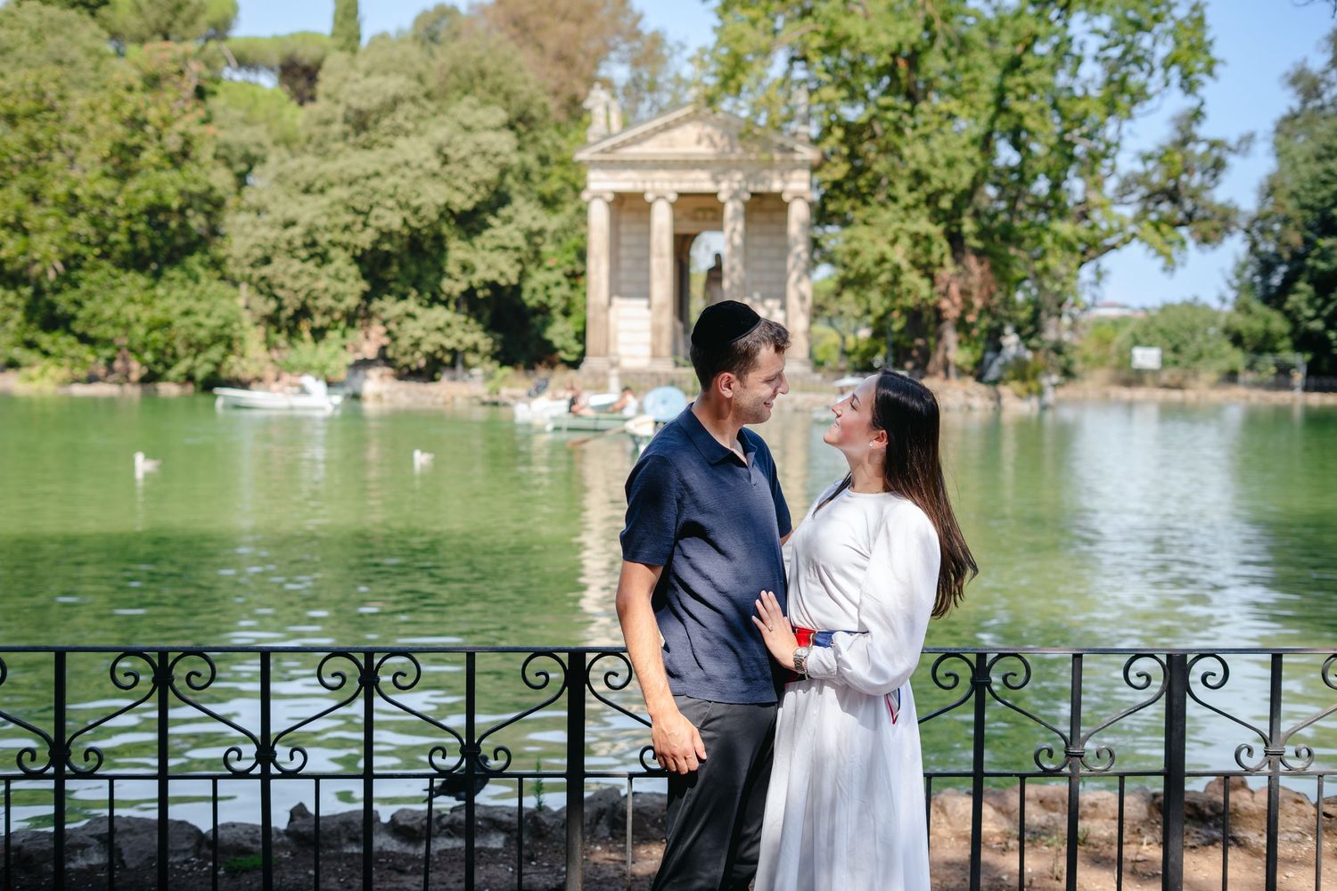 Couple embracing near a lake with a Roman temple and rowboats at Villa Borghese gardens in Rome.