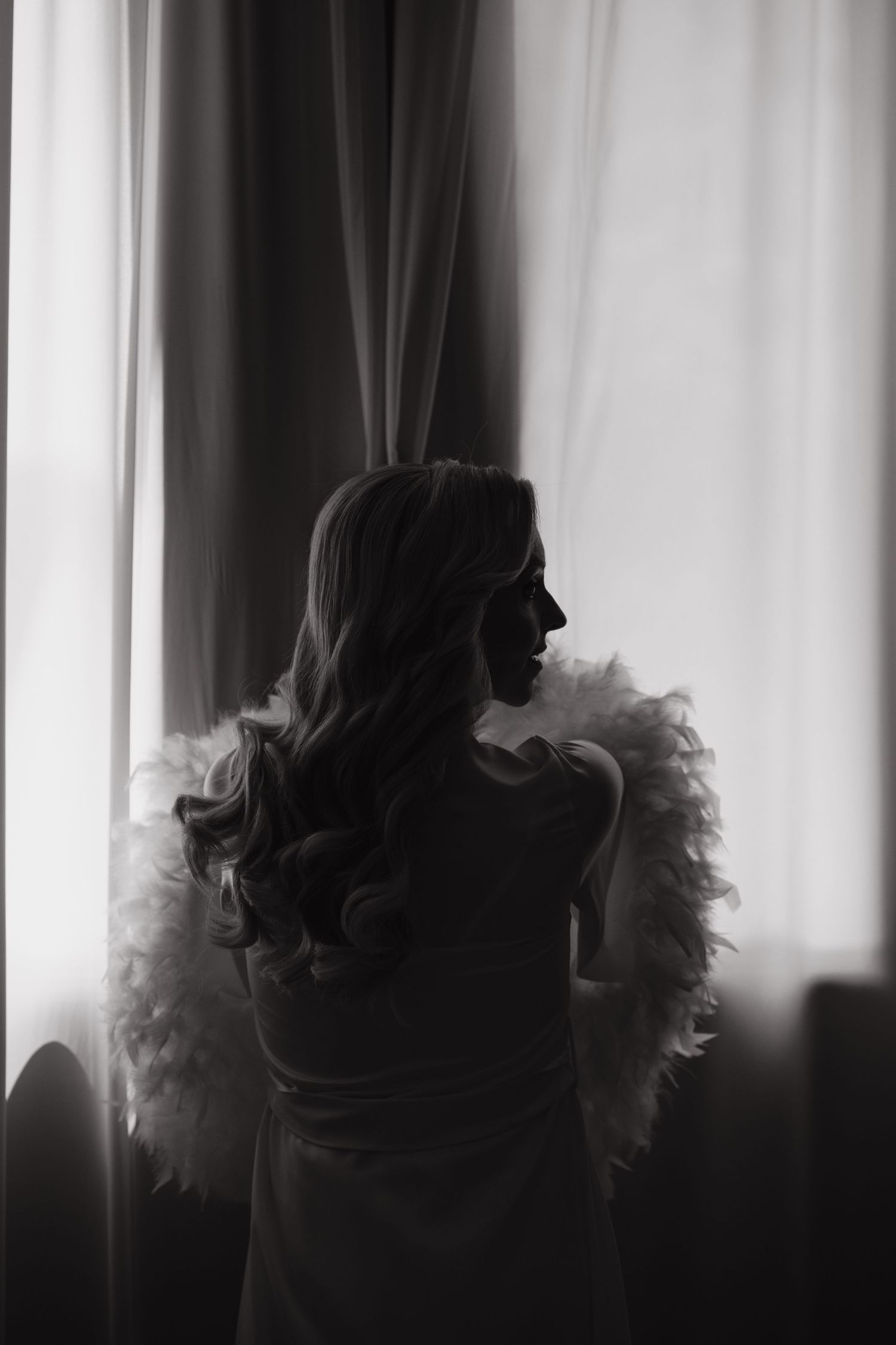 Black and white silhouette of a woman with long curly hair wearing a feather boa, standing by sheer curtains.
