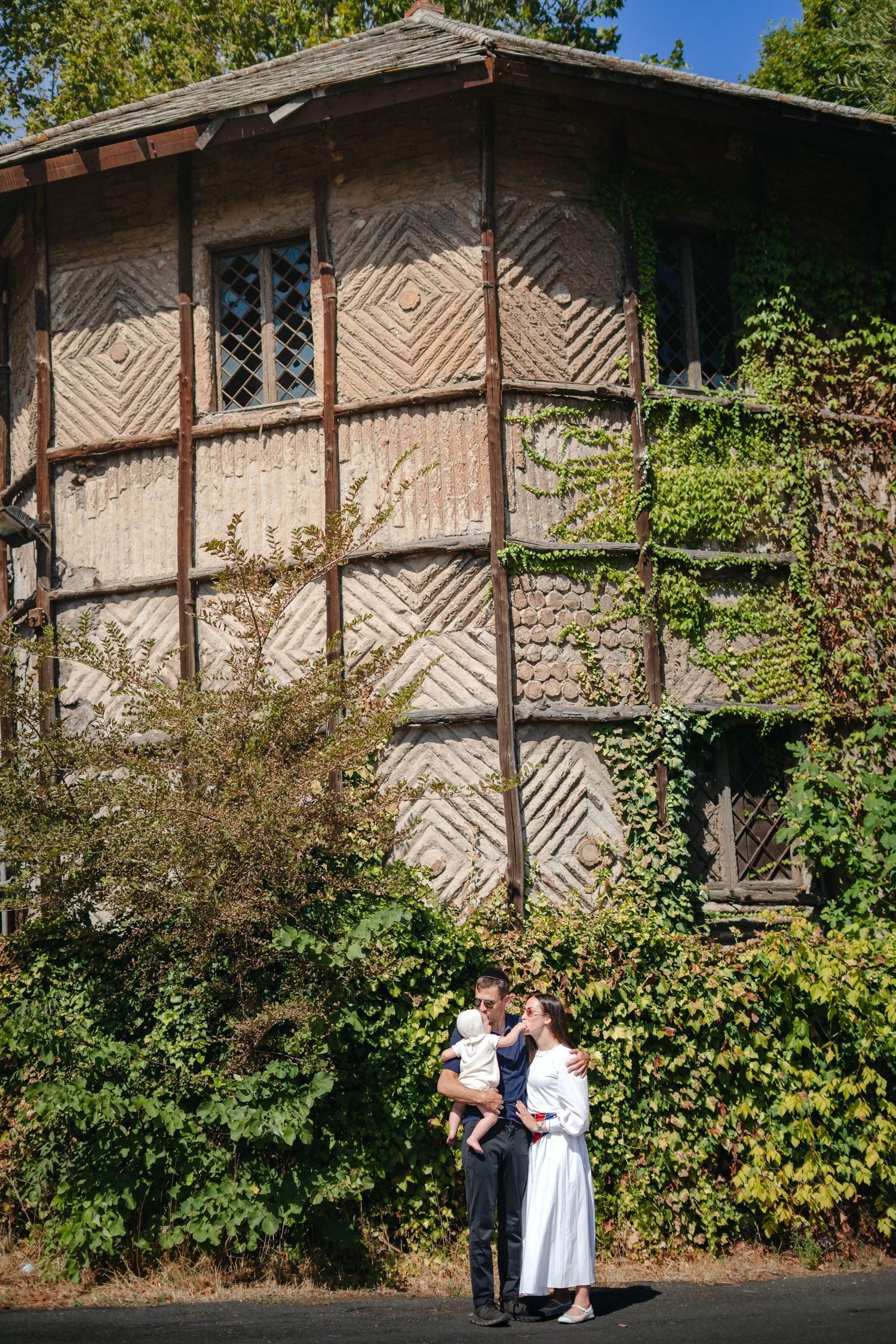 Family of three posing in front of a rustic half-timbered building covered in green ivy on a sunny day.