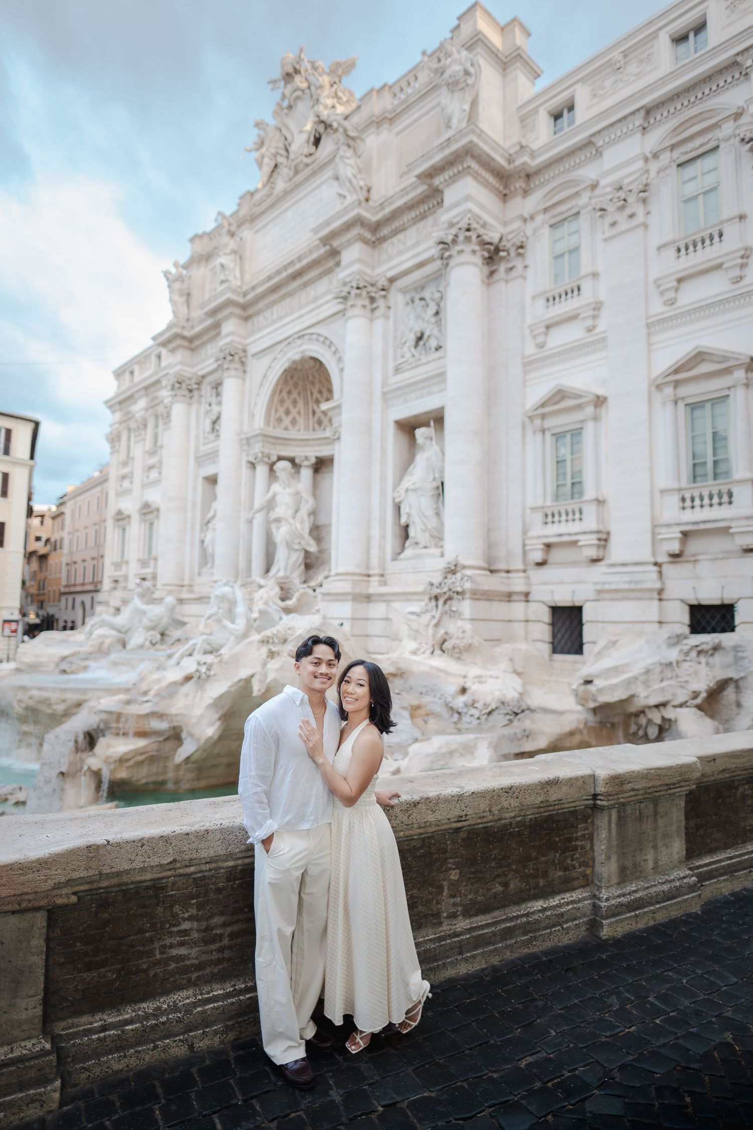 Couple in white outfits posing romantically in front of the iconic Trevi Fountain in Rome, Italy.