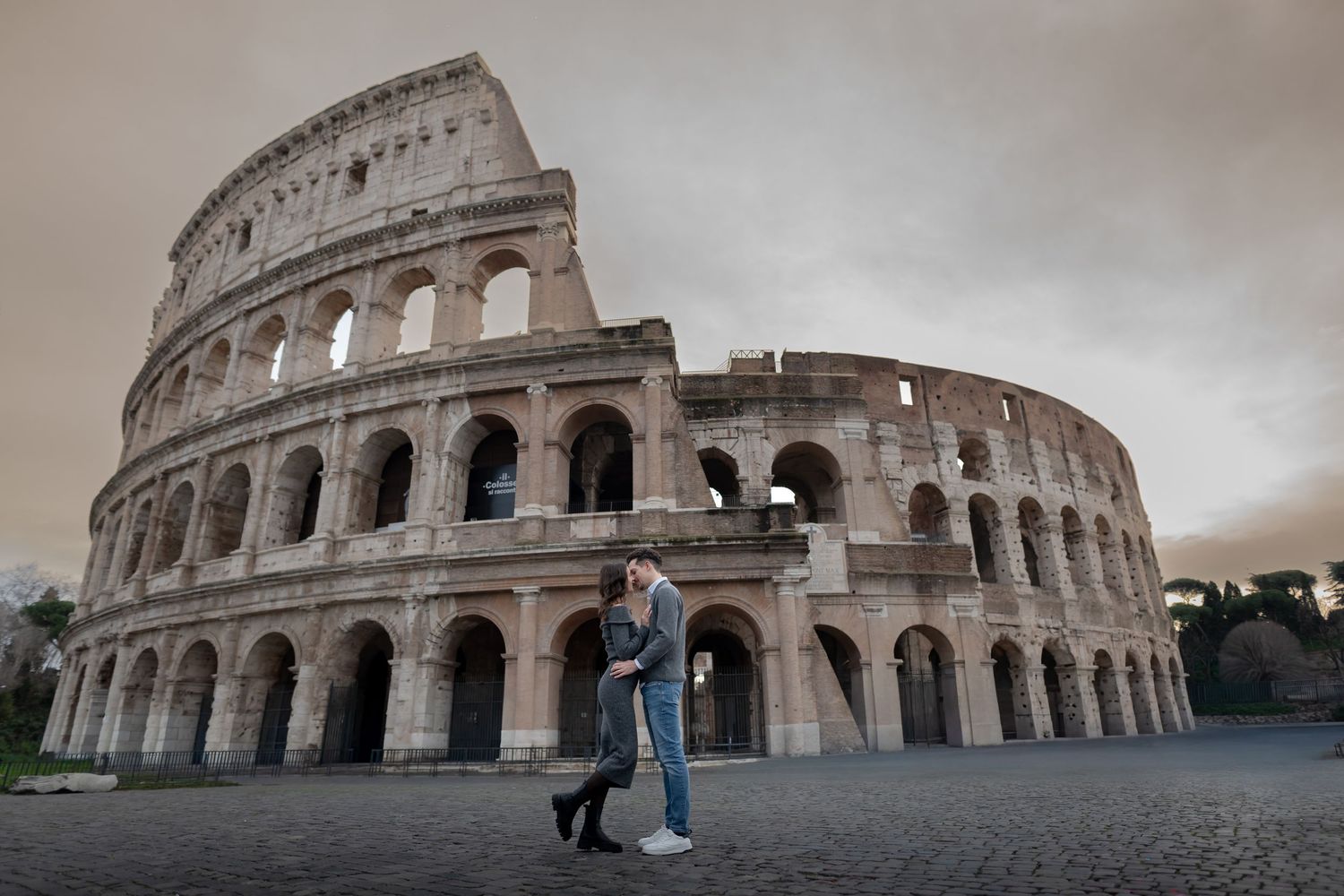 A couple embraces in front of the ancient Roman Colosseum under a dramatic cloudy sky in Rome, Italy.