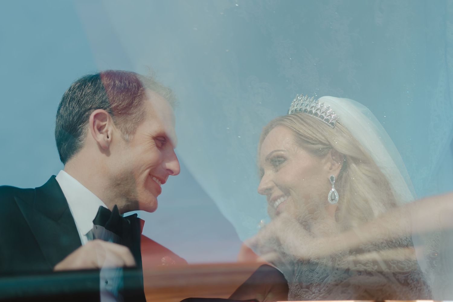 Bride and groom smiling at each other through a car window, bride wearing tiara and veil on wedding day.
