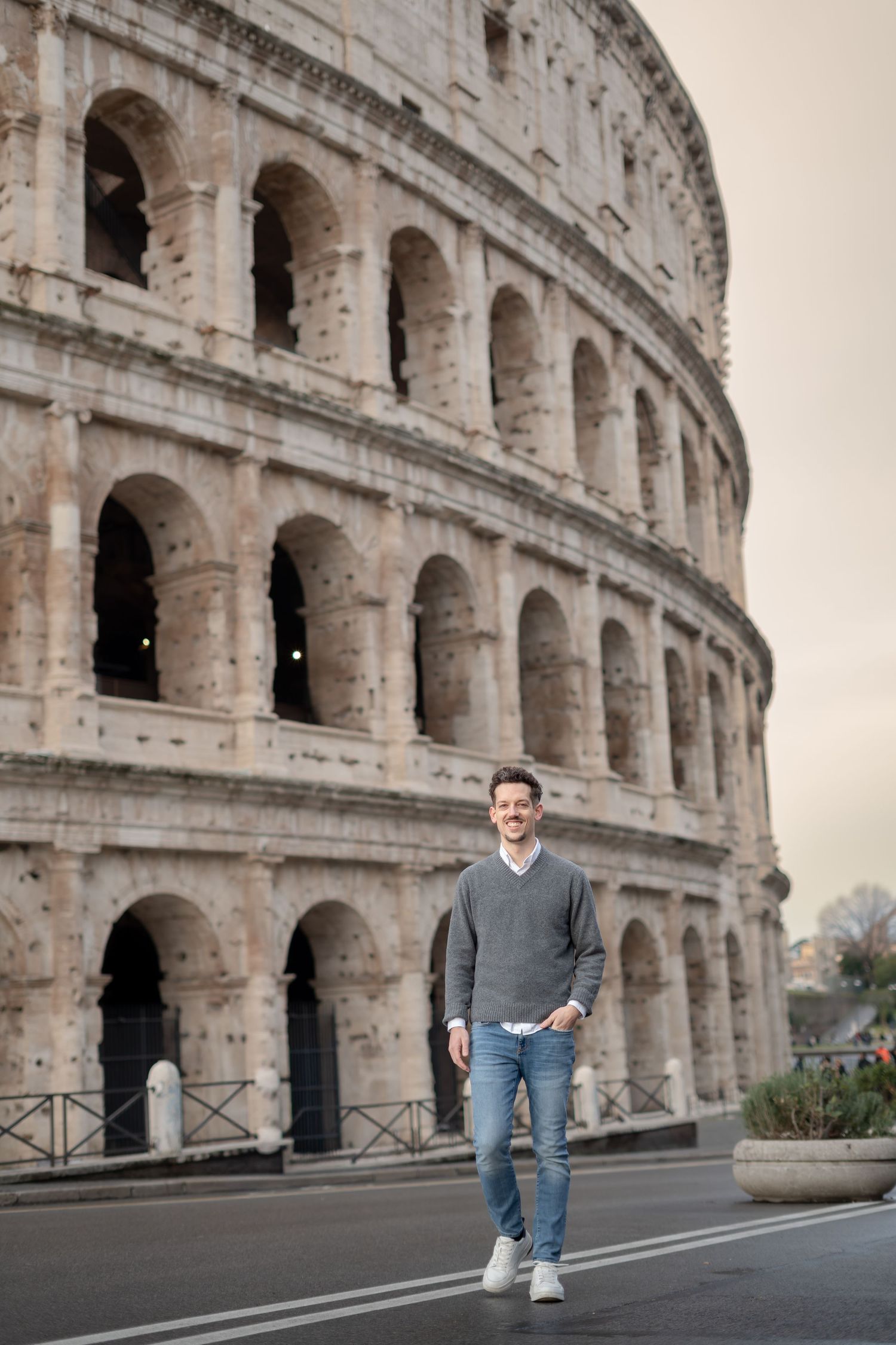 Smiling man in gray sweater and jeans walks in front of the historic Roman Colosseum in Rome, Italy.