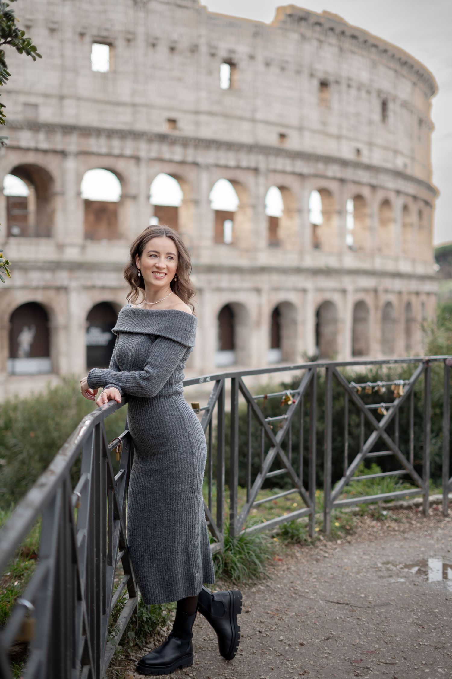 Woman in gray off-shoulder knit dress posing by metal railing with Rome's Colosseum in background.