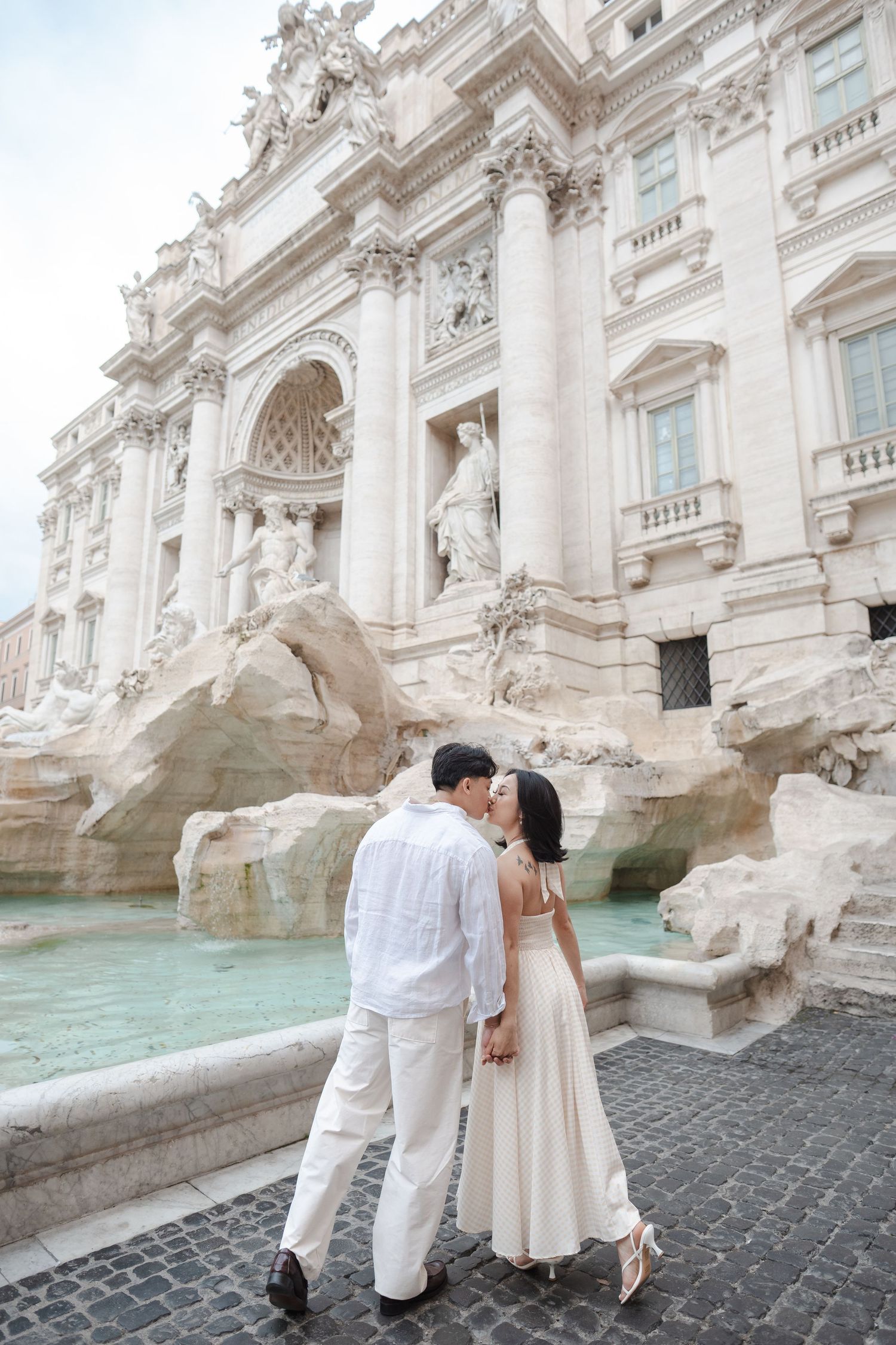 Couple kissing in front of the iconic Trevi Fountain in Rome, Italy, dressed in elegant white outfits.