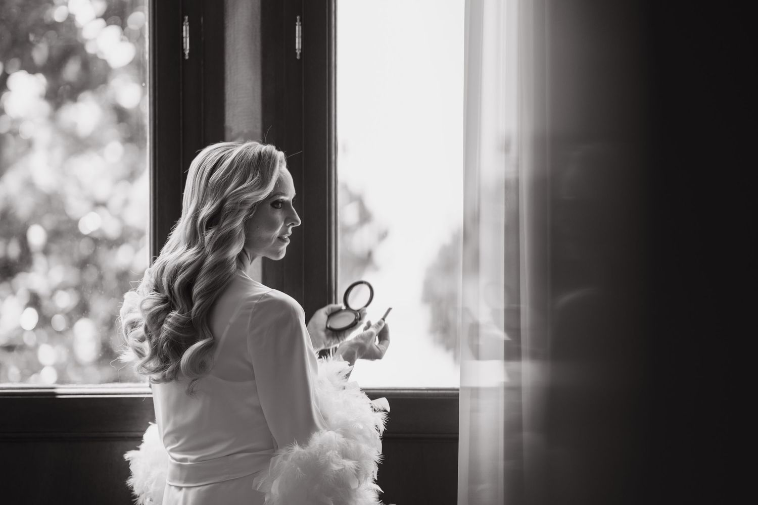 Elegant black and white portrait of a bride holding a compact mirror near a sunlit window.