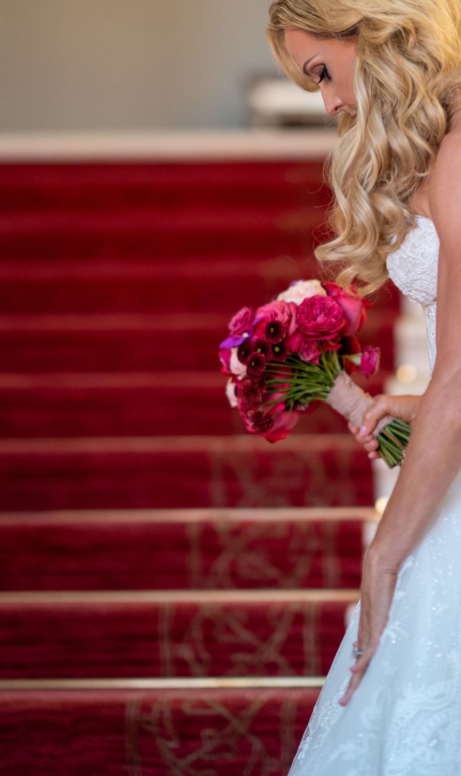 Blonde bride in white lace gown holds vibrant pink bouquet on red carpeted staircase, looking down elegantly.
