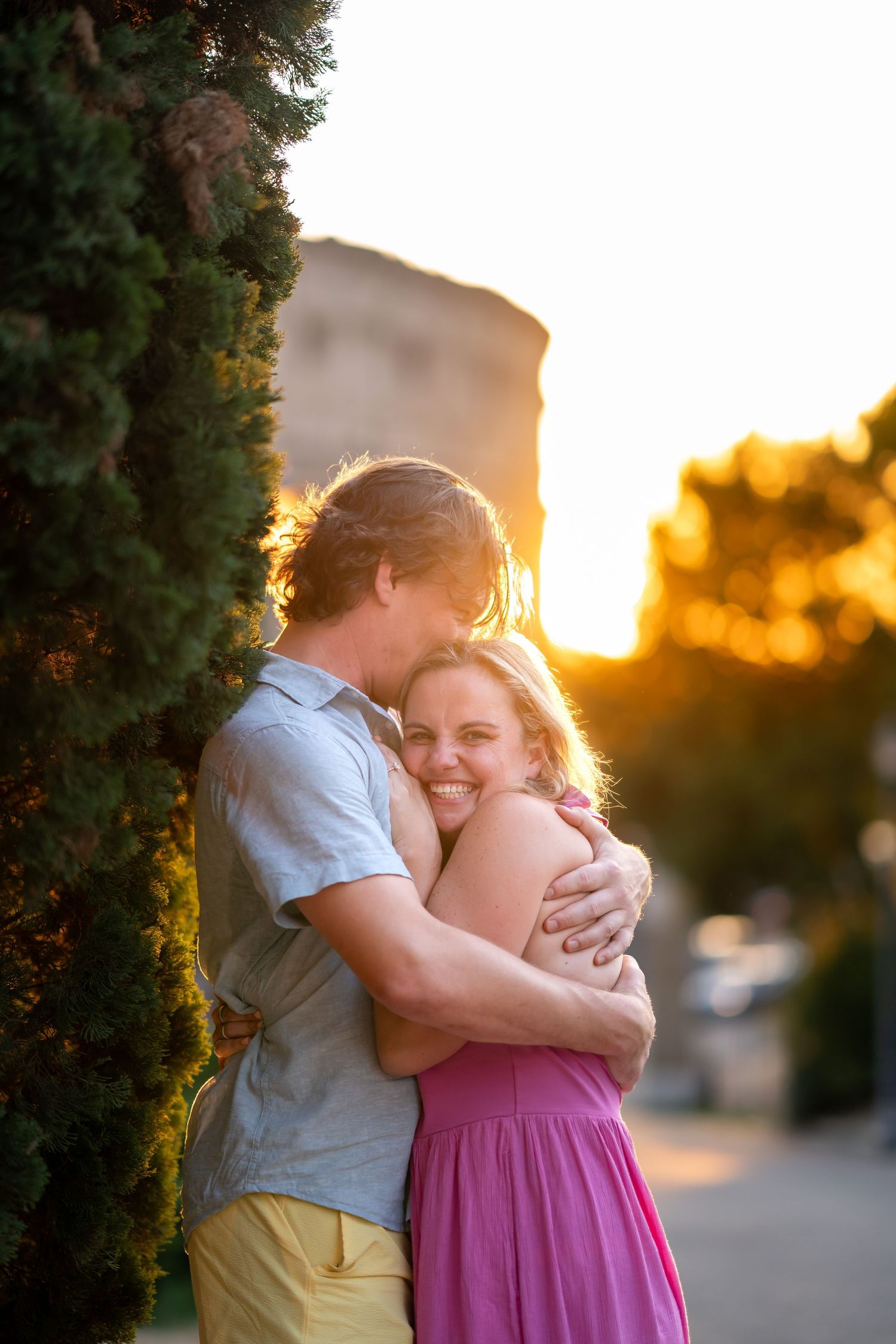 Couple embracing at sunset near cypress trees, woman in pink dress smiling joyfully, golden hour glow in background.