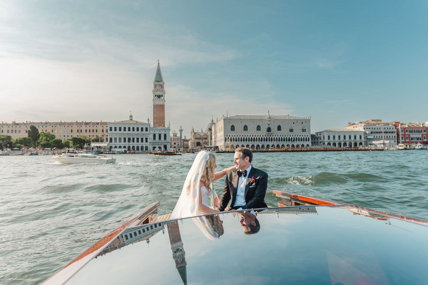 Bride and groom pose romantically on a wooden boat with Venice skyline and Campanile behind them.
