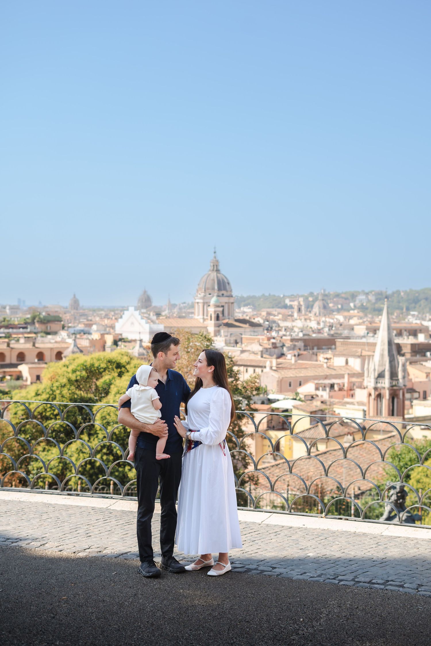 Young family with baby posing at a scenic Rome rooftop viewpoint with city skyline and domes in background.