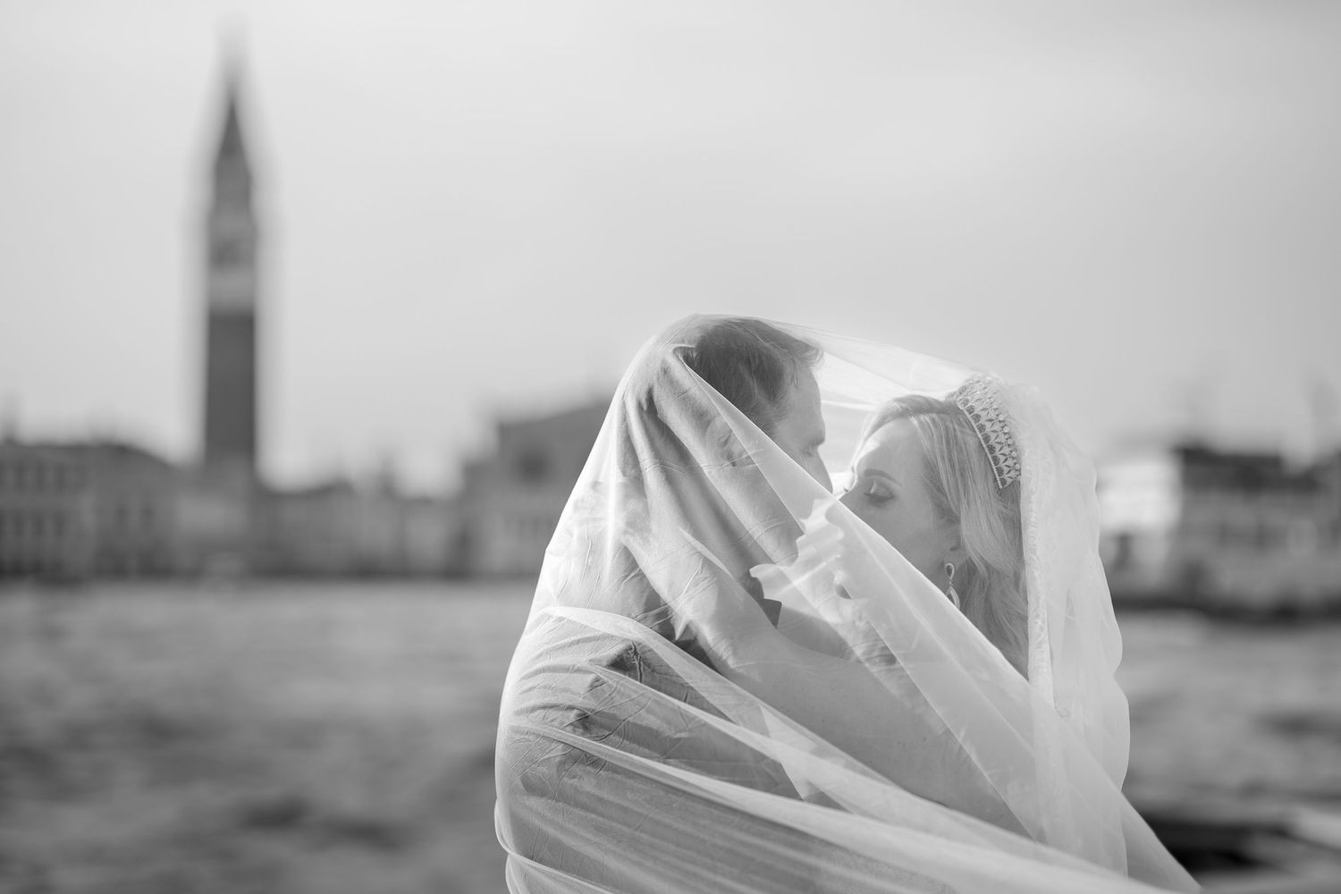 Black and white romantic photo of couple kissing under bride's veil with Venice skyline in background.