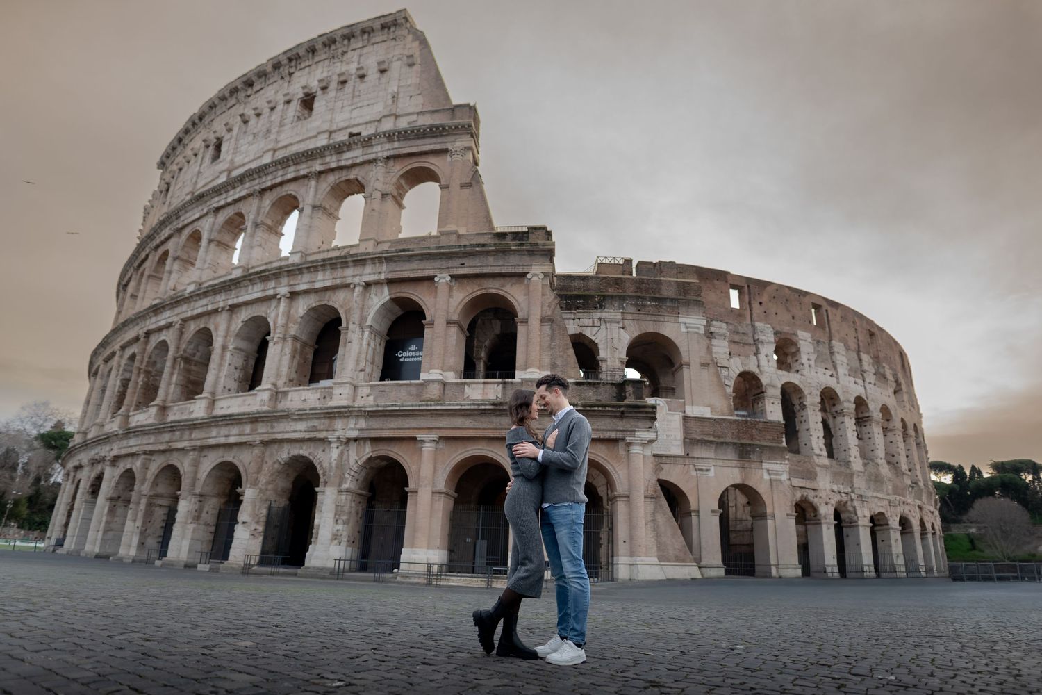 Couple sharing a romantic moment in front of the ancient Roman Colosseum under a dramatic cloudy sky.