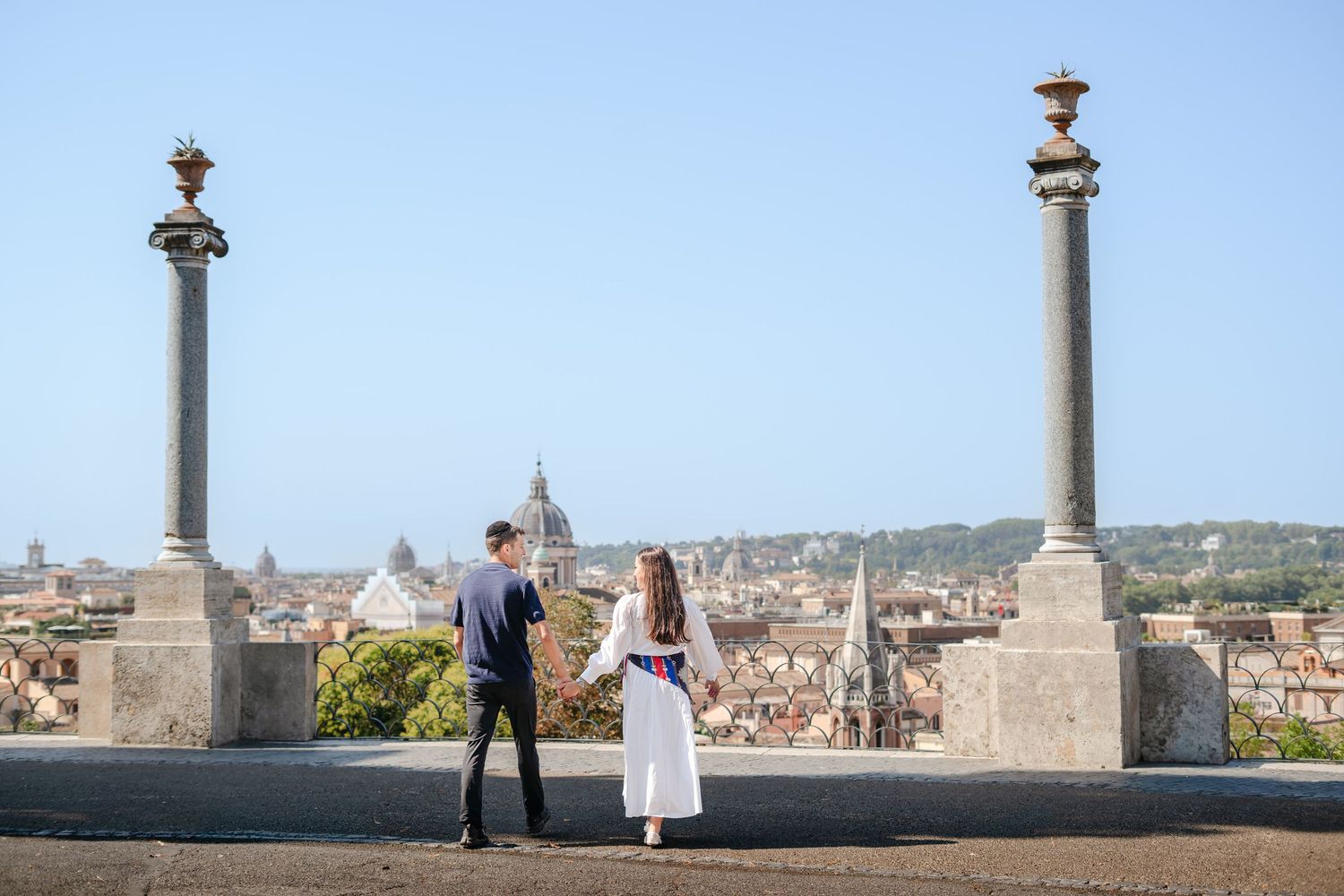 Couple holding hands on a rooftop terrace overlooking the Rome skyline with historic domes and columns.