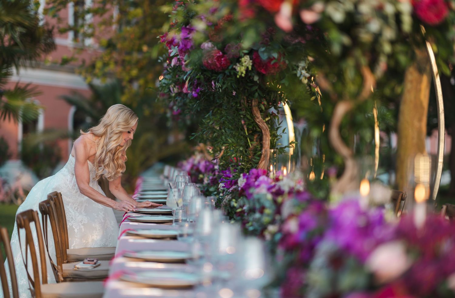 Bride in lace wedding gown arranging settings at elegantly decorated outdoor reception table with purple florals.