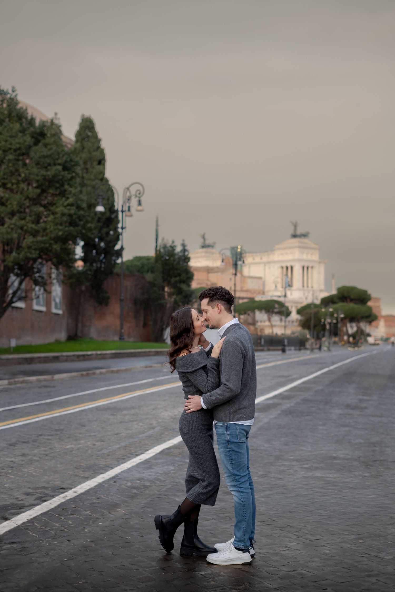 Romantic couple embracing and kissing on a wet street in Rome with the Altare della Patria monument in the background.