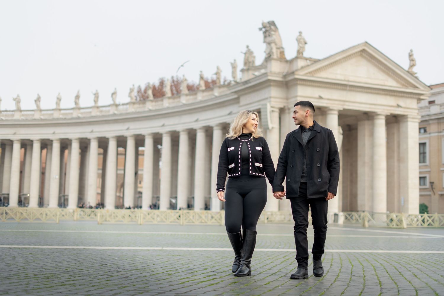 Couple holding hands walking in St. Peter's Square, Vatican City, dressed in black outfits with colonnade backdrop.