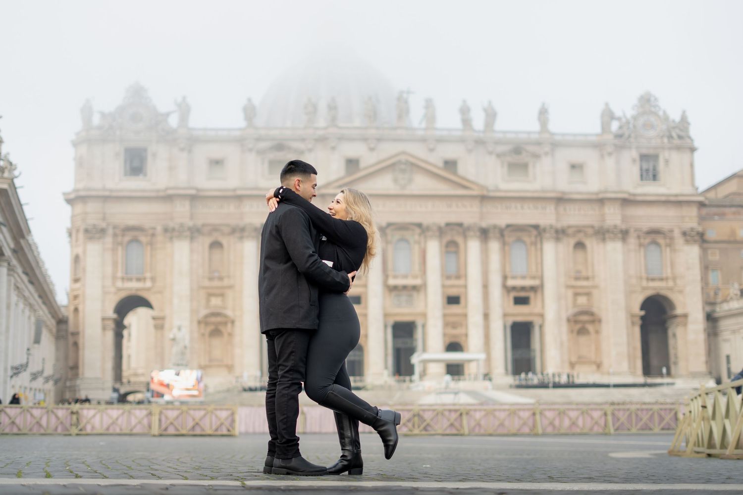 Couple embracing romantically in St. Peter's Square, Vatican City, on a misty day with the basilica in the background.