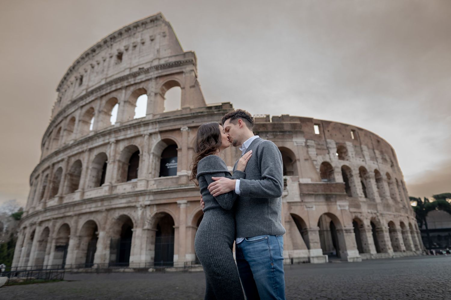 Couple kissing romantically in front of the ancient Colosseum in Rome, Italy, under a dramatic cloudy sky.