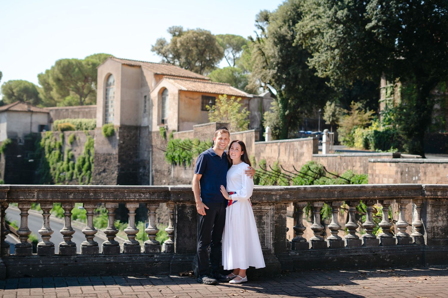 Couple posing together on a stone balustrade terrace with lush greenery and historic Roman architecture in the background.