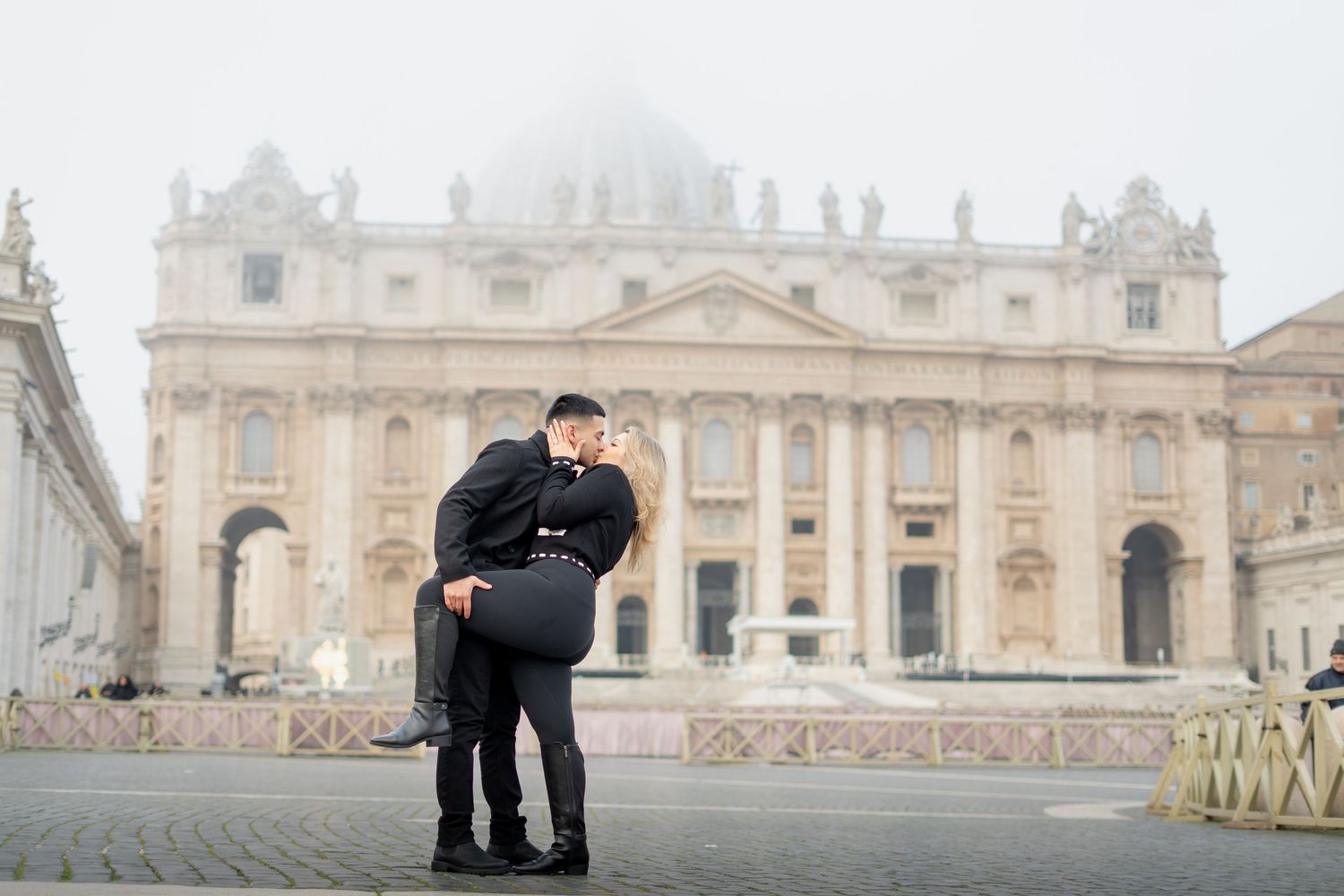 Couple sharing a romantic kiss in St. Peter's Square, Vatican City, on a misty day.