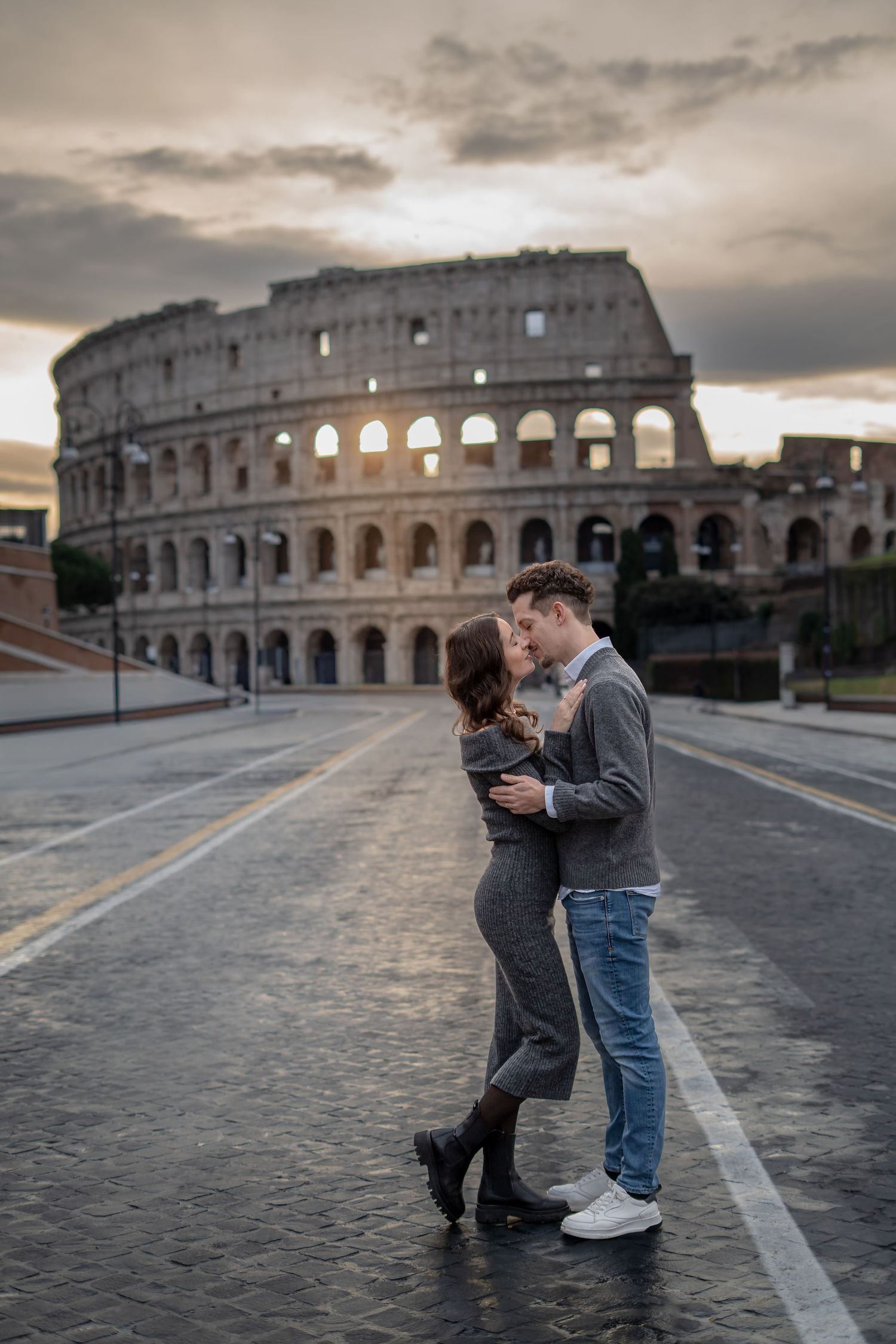 Romantic couple kissing in front of the Colosseum in Rome at sunset on an empty street.