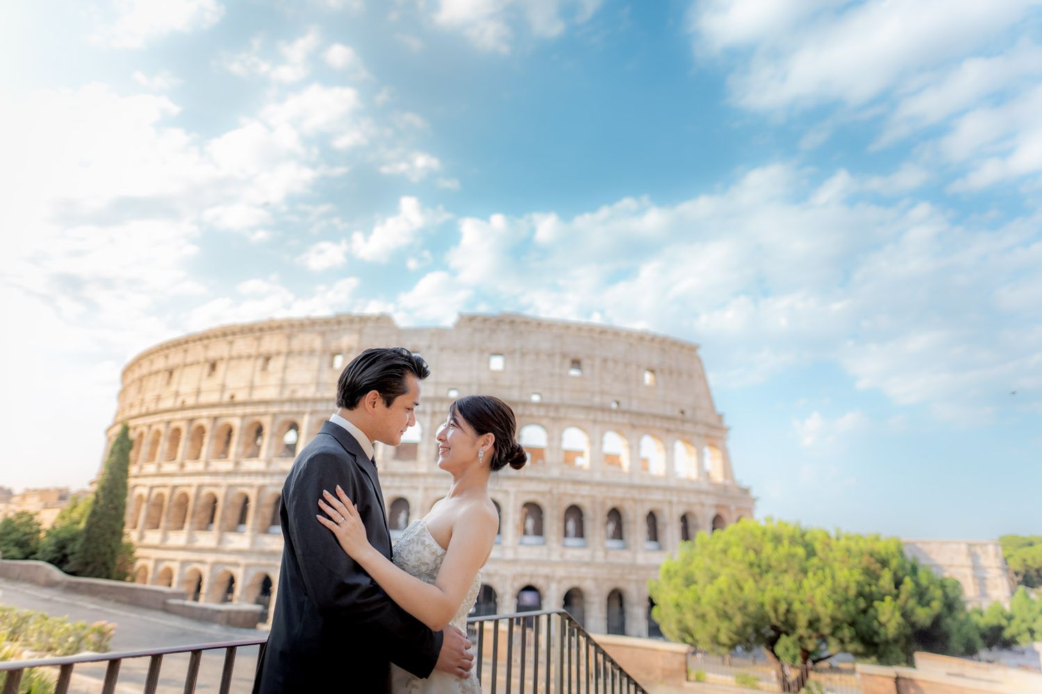 Couple in wedding attire embracing in front of the Colosseum in Rome, Italy, under a bright blue sky.