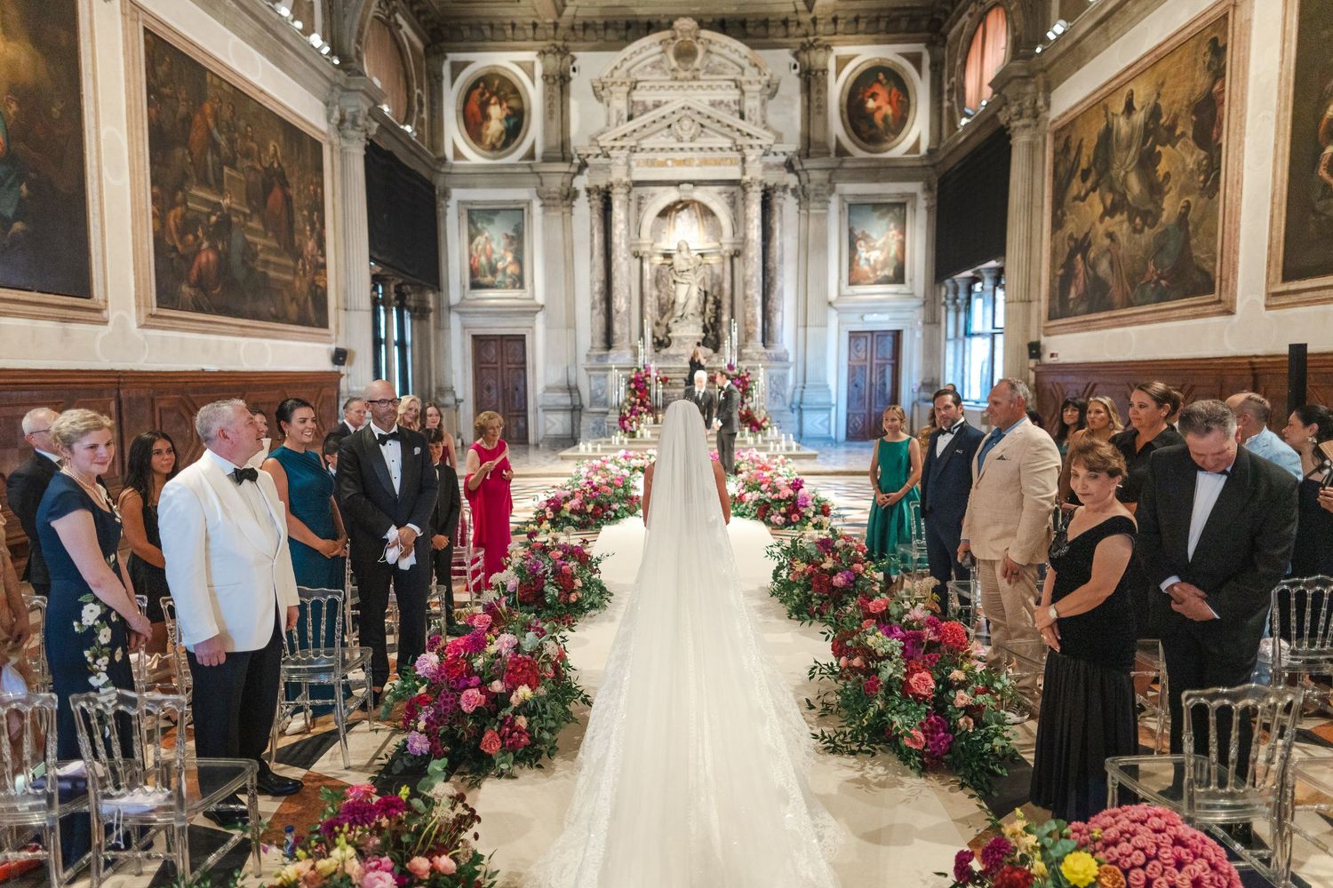 Bride walks down flower-lined aisle in ornate Venetian church surrounded by wedding guests.