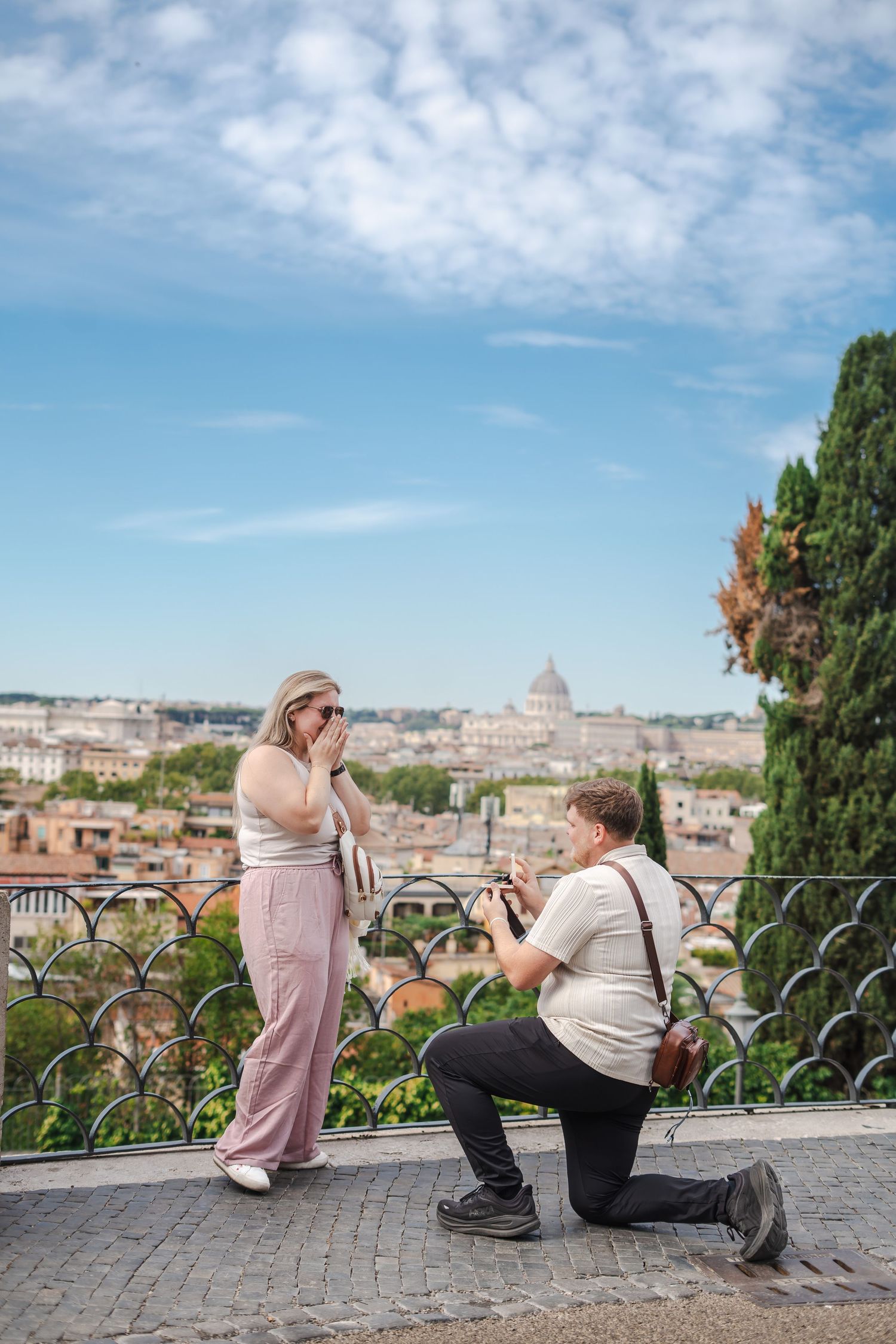 Man proposes on one knee to surprised woman at scenic Rome overlook with St. Peter's Basilica in background.