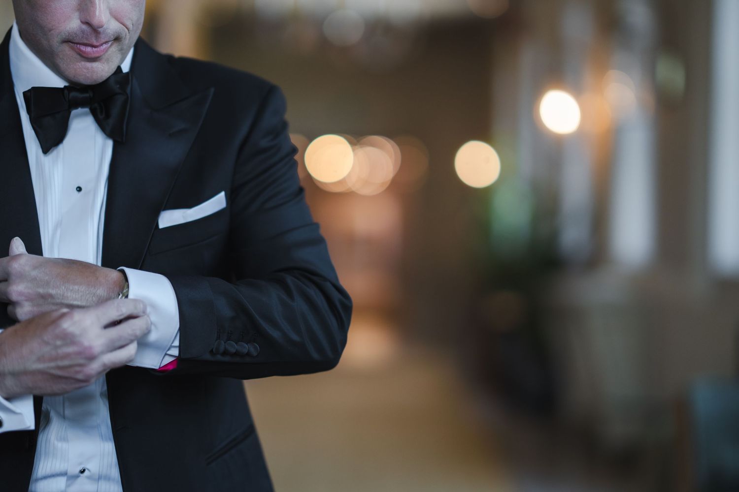 Groom in black tuxedo and bow tie adjusting his cufflinks with bokeh lights in the background.