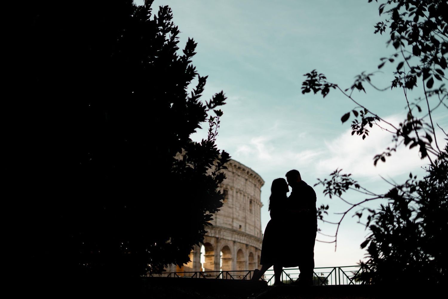 Silhouette of a romantic couple kissing near the Colosseum in Rome at dusk with trees framing the scene.
