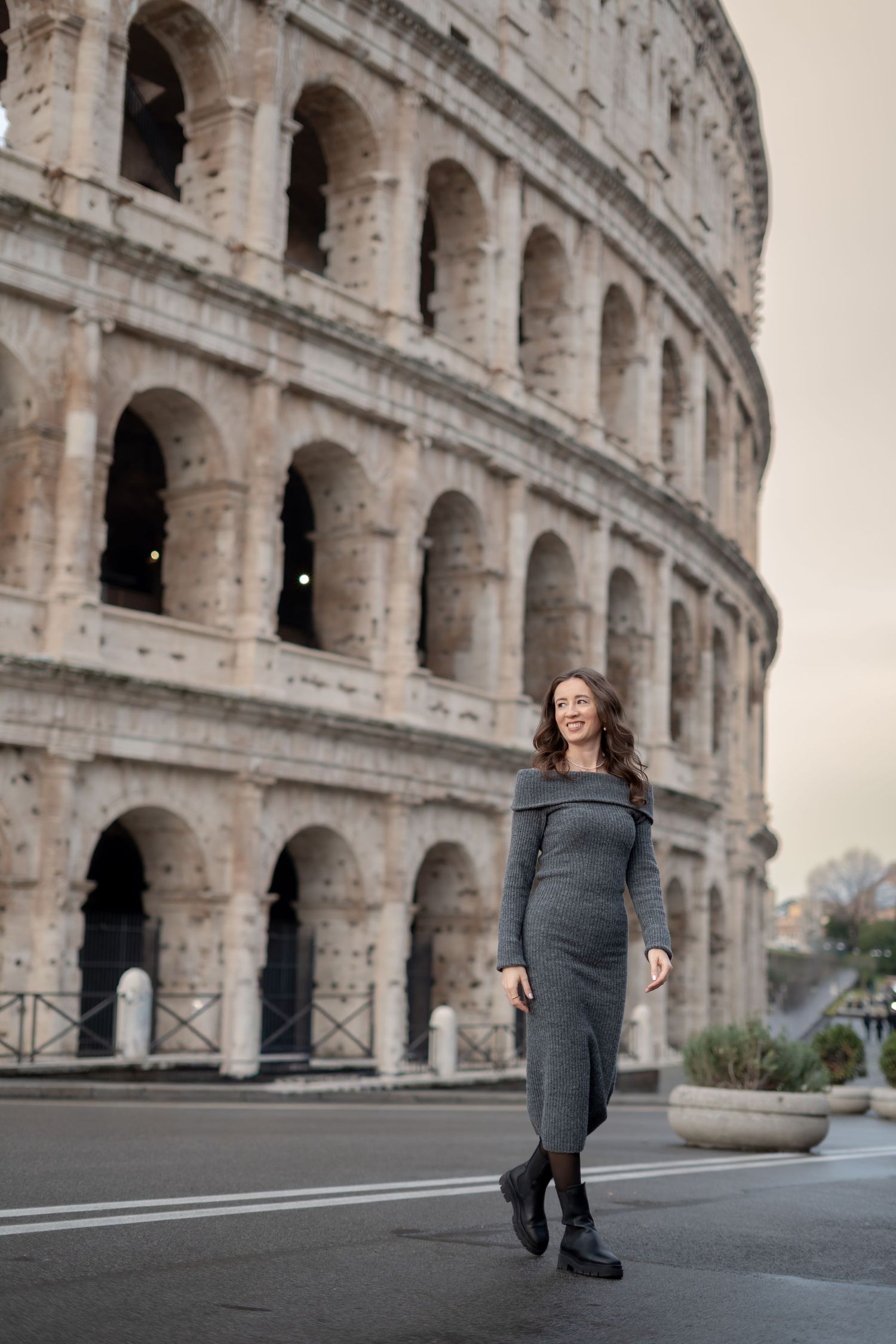 A woman in a gray off-shoulder dress walks in front of the iconic Colosseum in Rome, Italy.