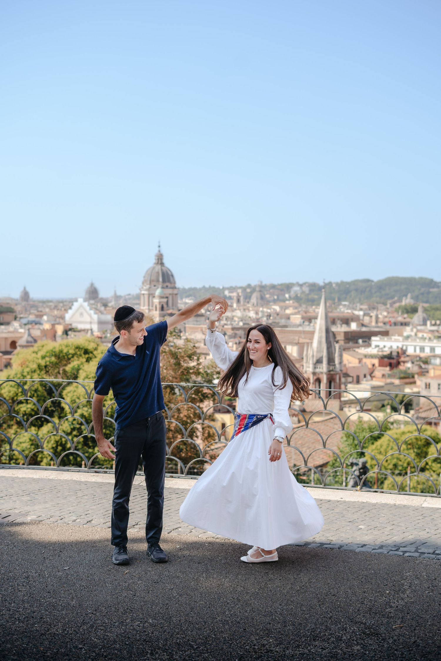 Couple dancing on a terrace overlooking Rome's skyline with domes and rooftops on a clear sunny day.