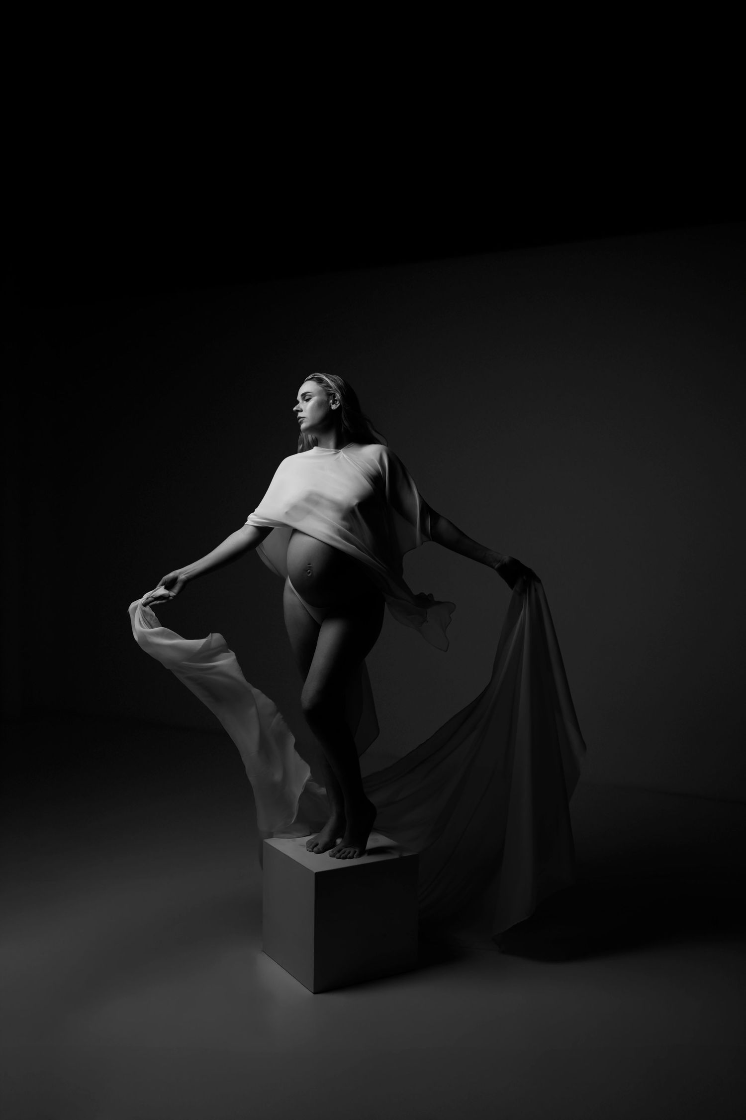 Black and white maternity photo of a pregnant woman draped in flowing fabric, standing on a cube in a dramatic studio setting.