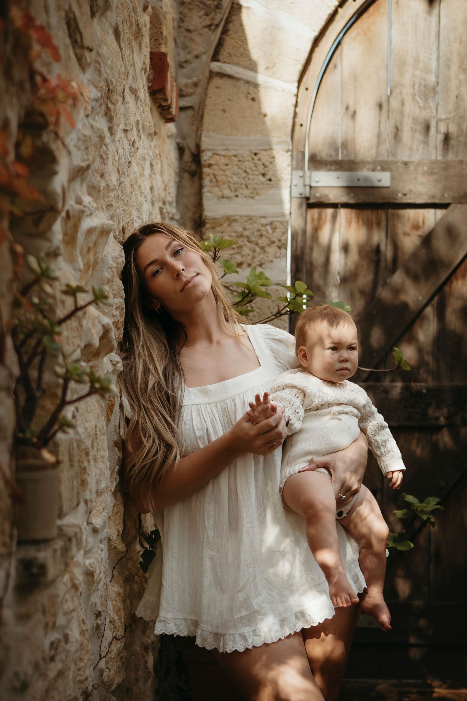 Woman in white dress holds baby against rustic stone wall with wooden door and green vines.