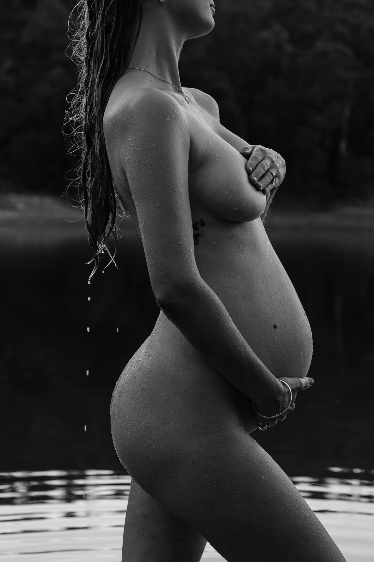 Black and white artistic maternity photo of pregnant woman standing near water with wet hair.