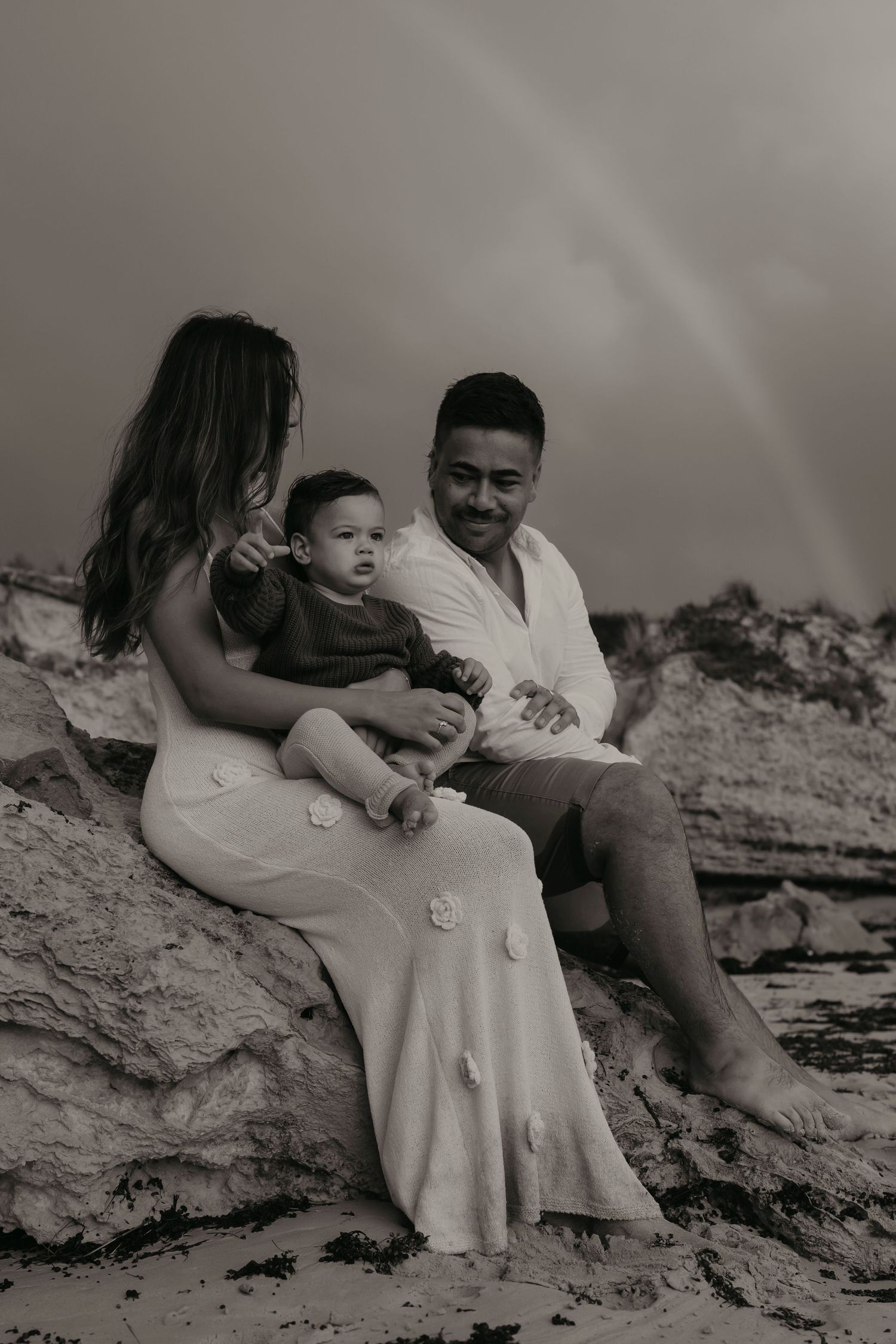 A family sits together on rocky beach terrain in a black and white portrait as they share an intimate moment.