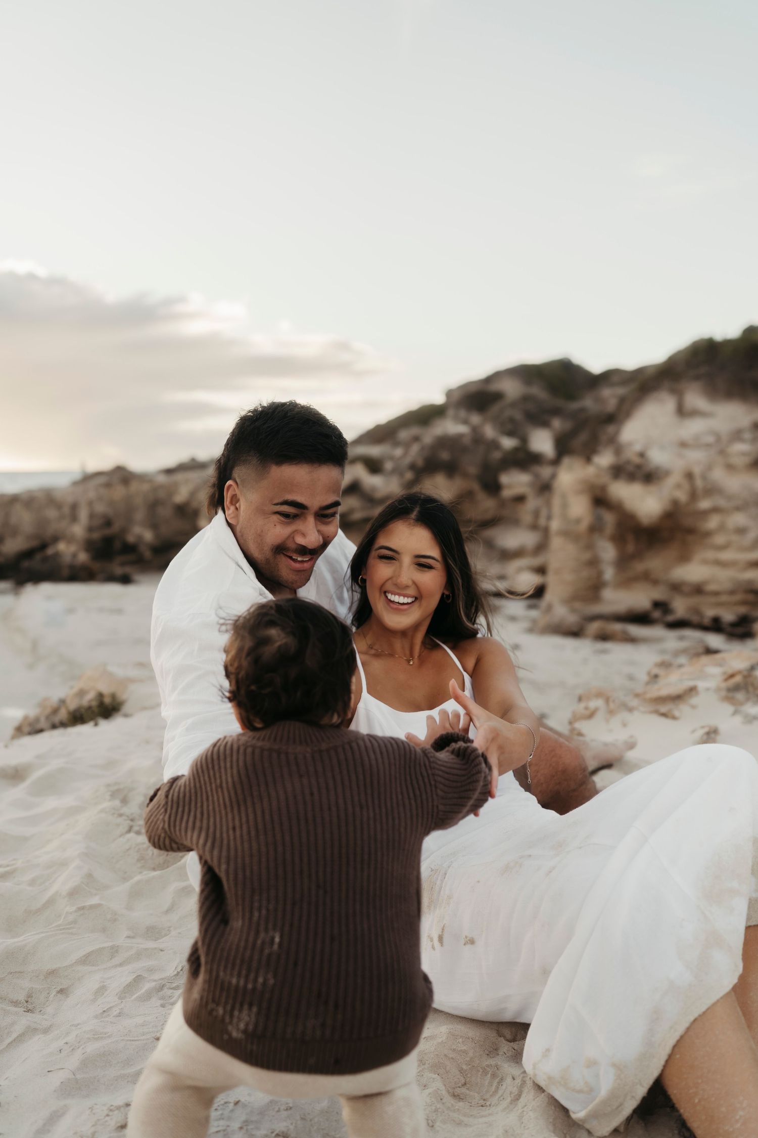 A joyful couple in wedding attire sharing a candid moment on a beach with rocky cliffs in the background.