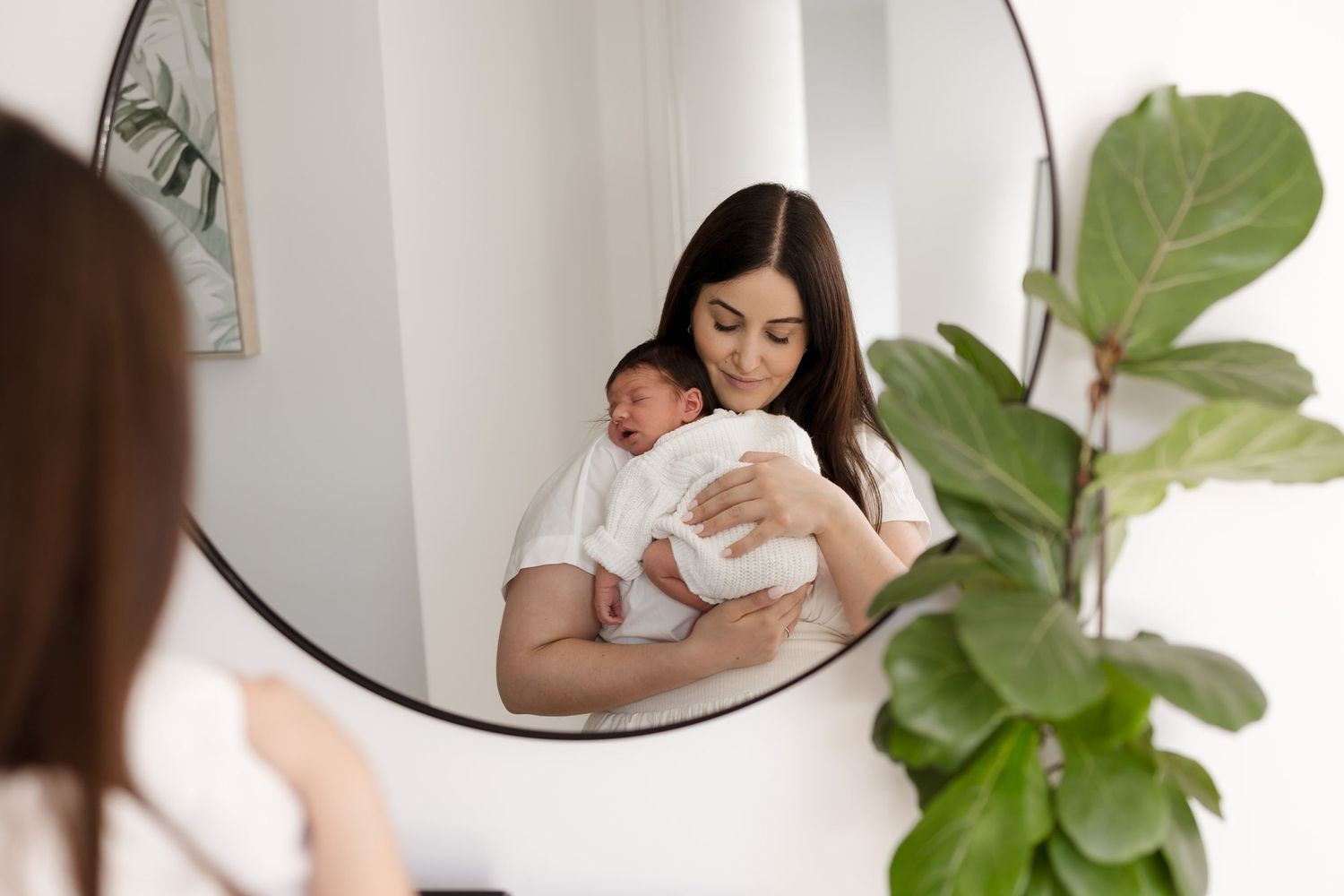 Mirror reflection shows mother holding newborn baby with houseplant in foreground. Meg Rose Photos Perth WA.