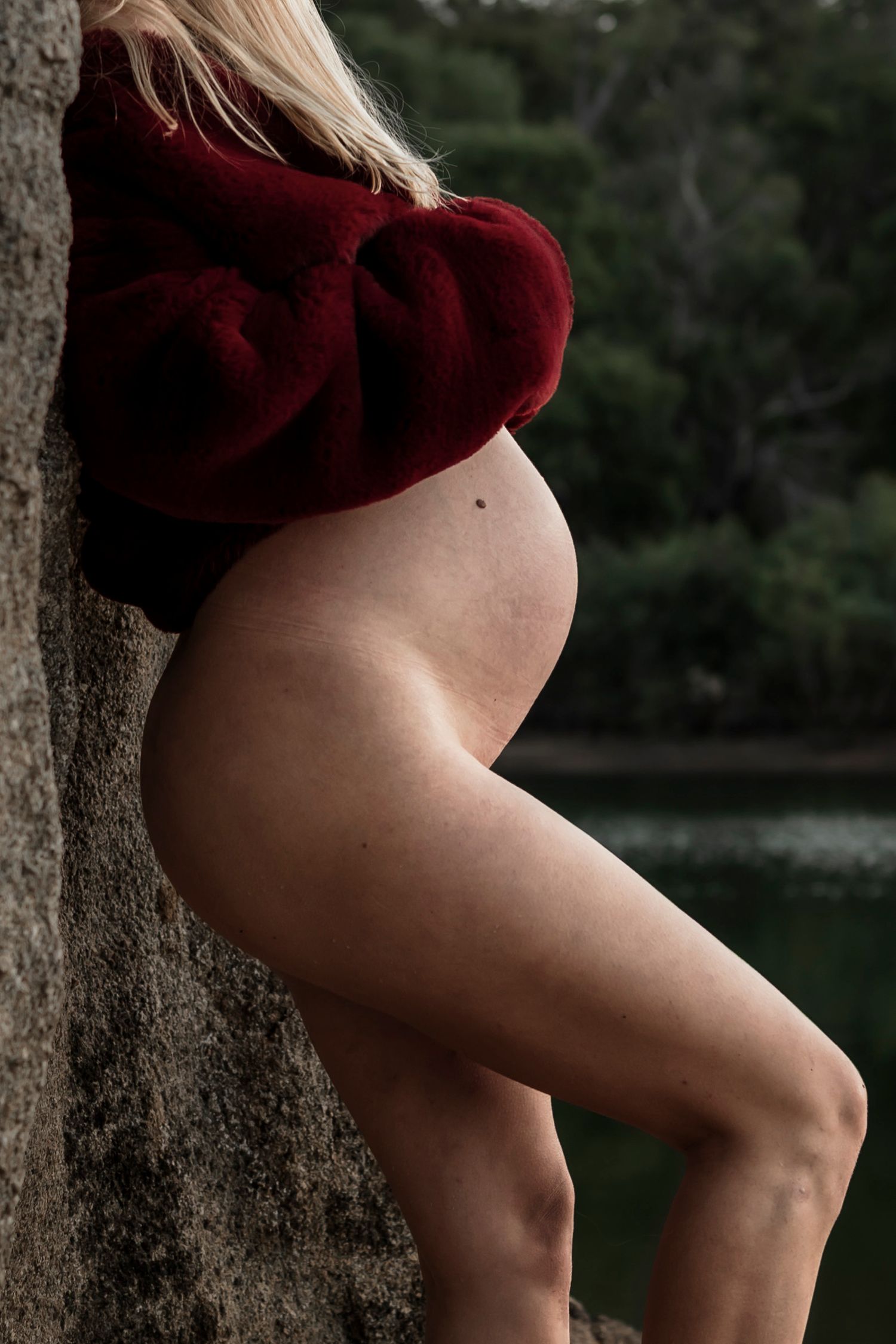 Silhouette of a pregnant figure in red sweater leaning against stone by water.