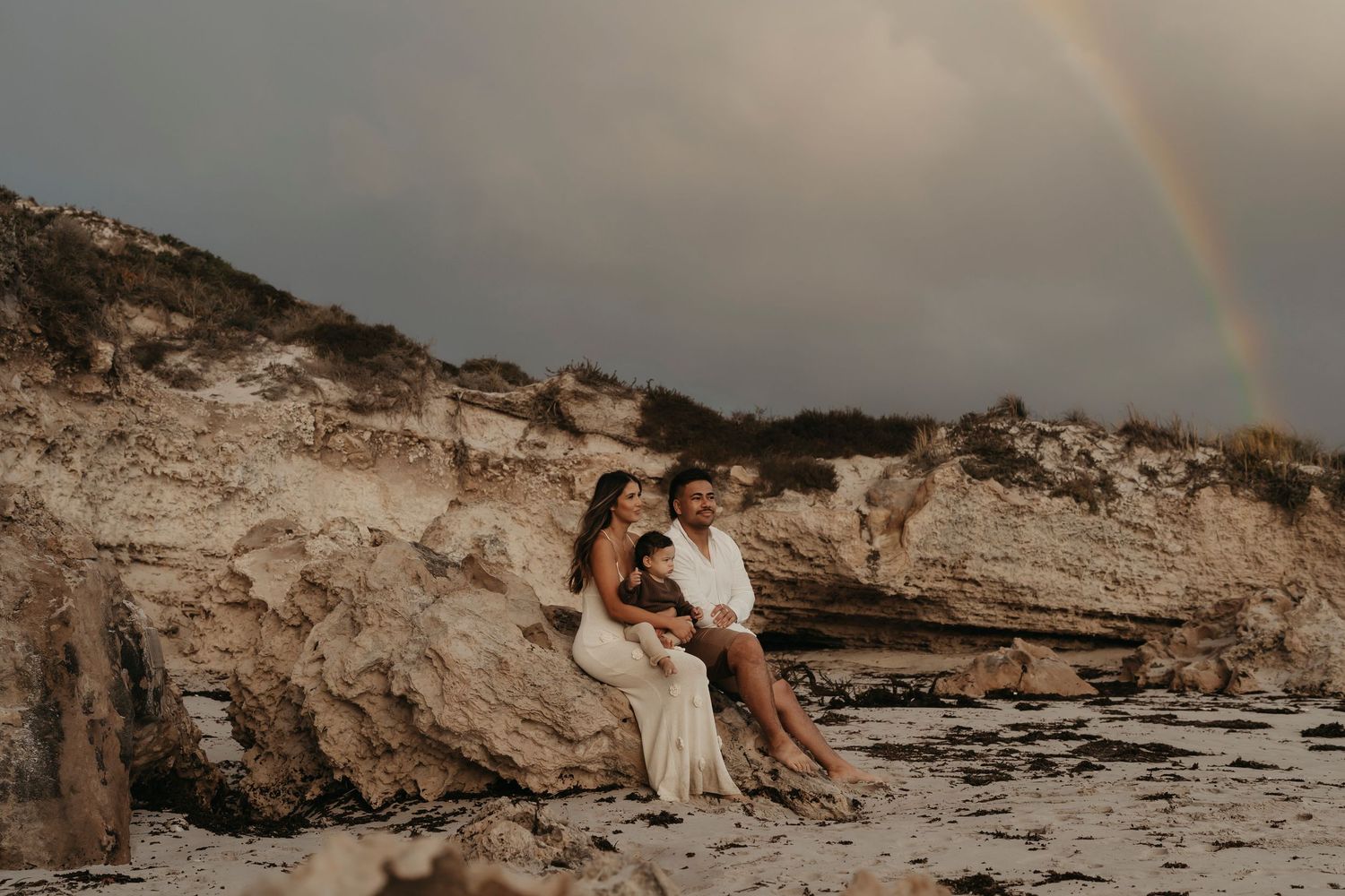 Couple sitting on rocky coastal cliffs with a rainbow appearing in the dramatic sky.