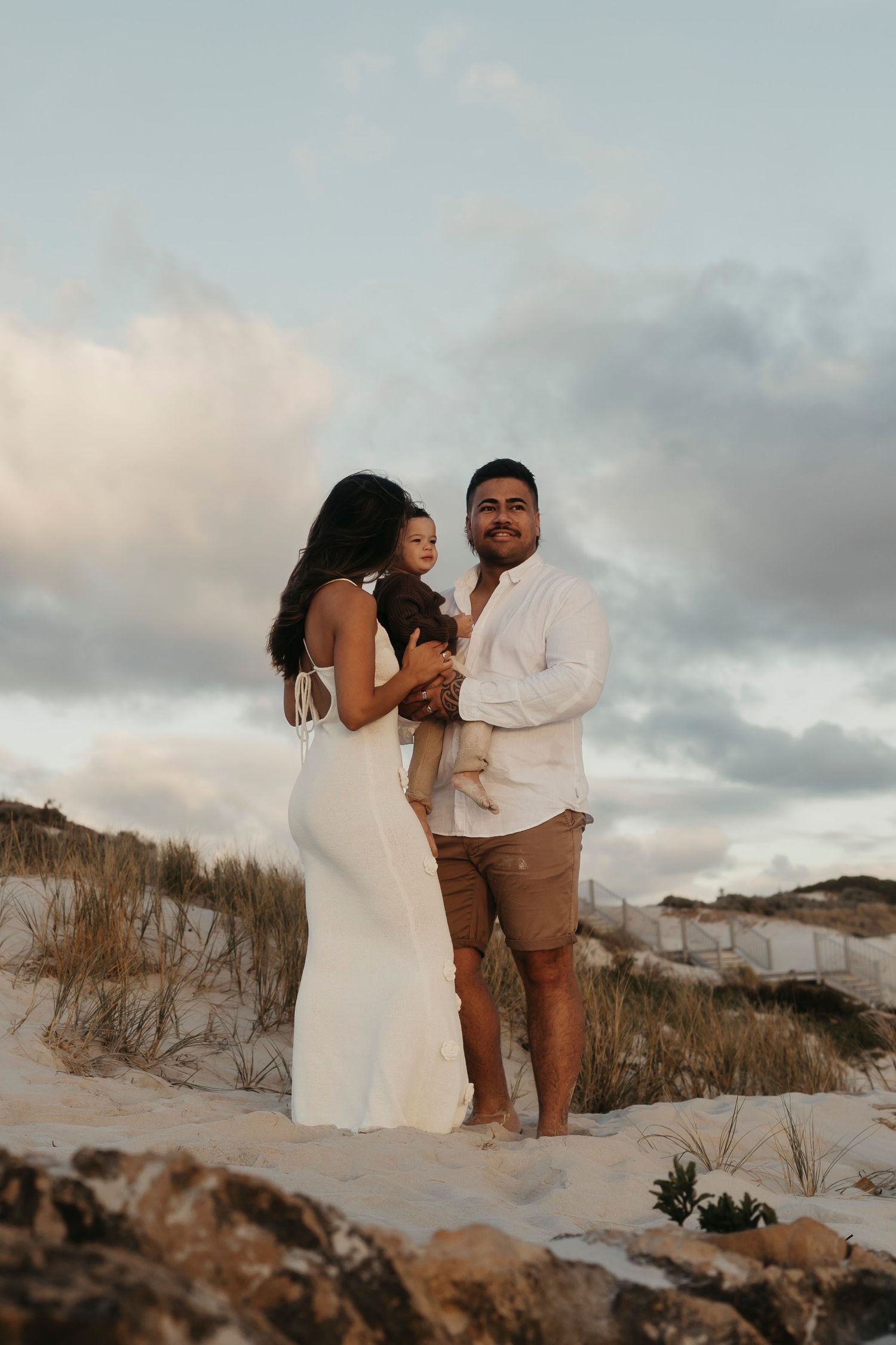 A couple standing on sandy dunes at sunset wearing light-colored outfits and smiling.