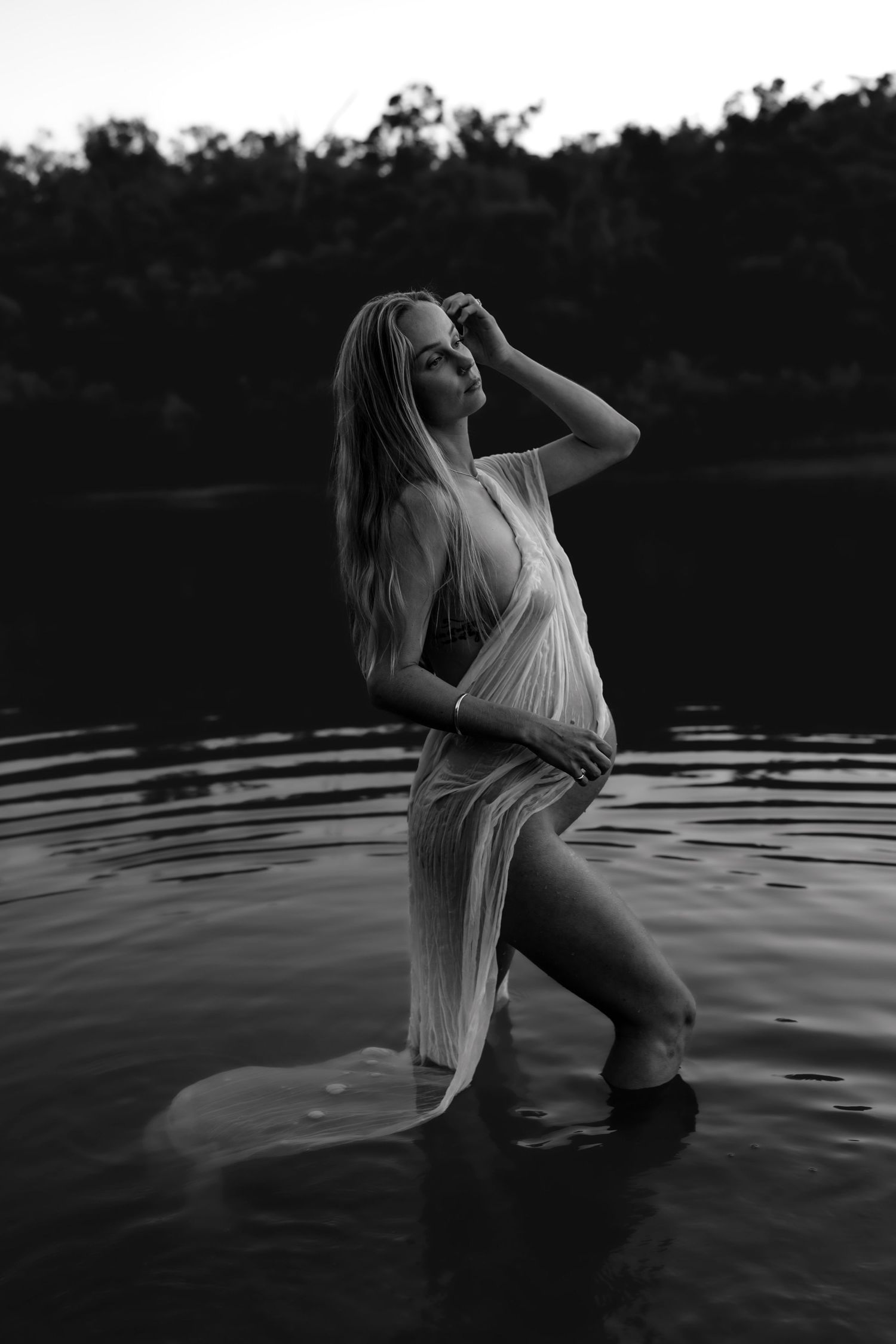 Artistic black and white maternity photo of an expectant mother standing in water wearing a sheer white dress.