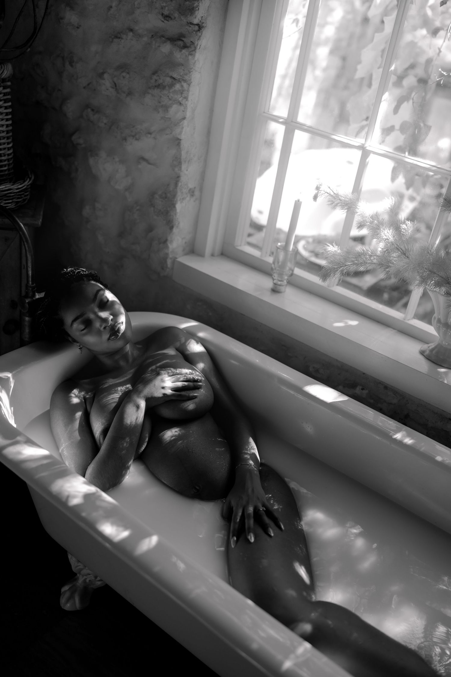 A woman reclines in a vintage clawfoot bathtub by a sunlit window in a black and white photo.