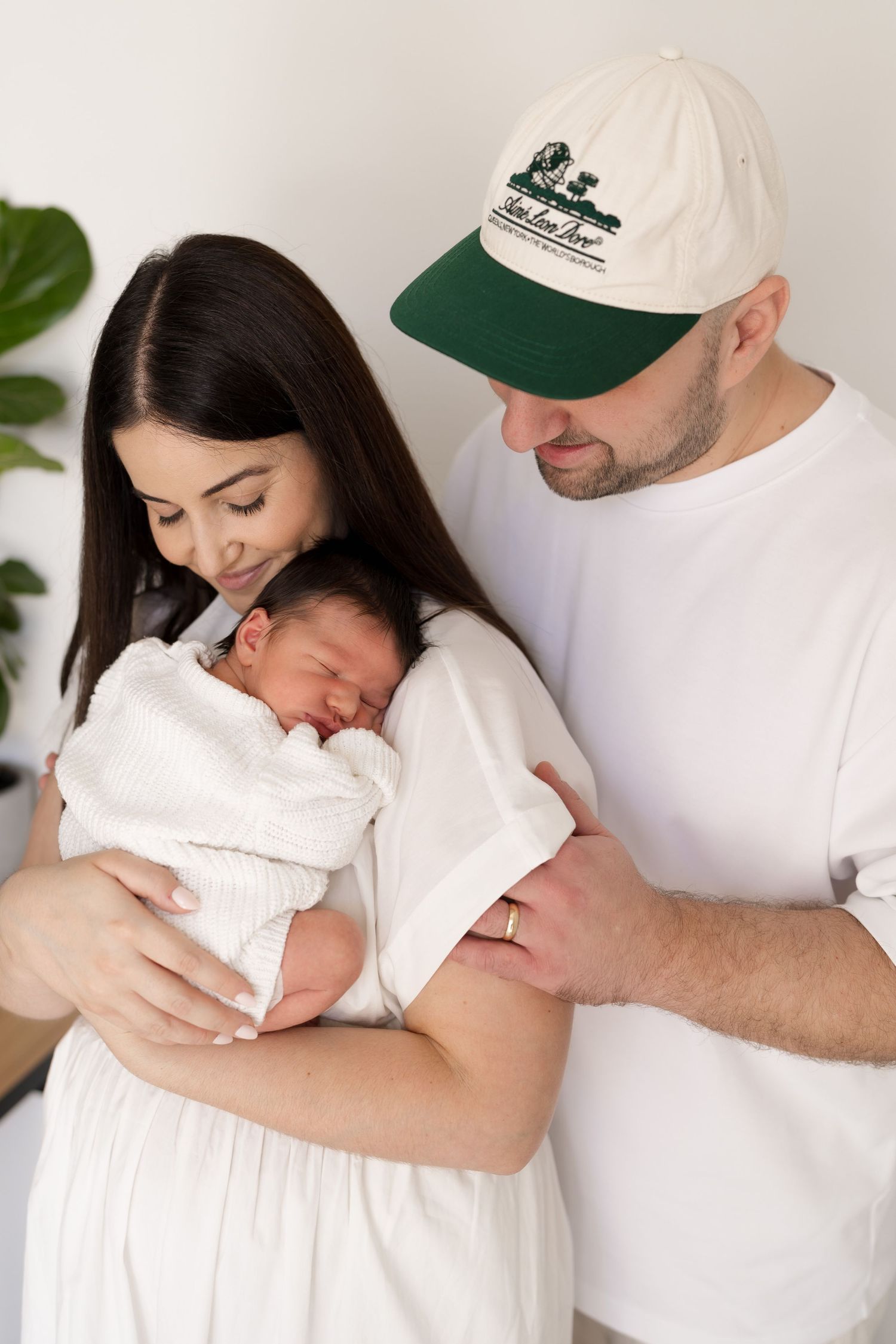 Family portrait with parents in white clothing holding sleeping newborn near green plant. Meg Rose Photos Perth WA.
