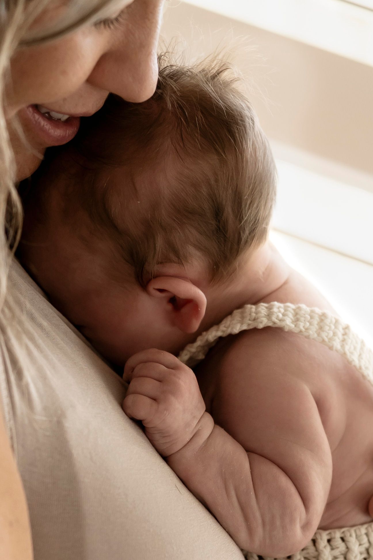 A mother tenderly holds her newborn baby close, the infant dressed in a knit romper, in a soft natural light setting.