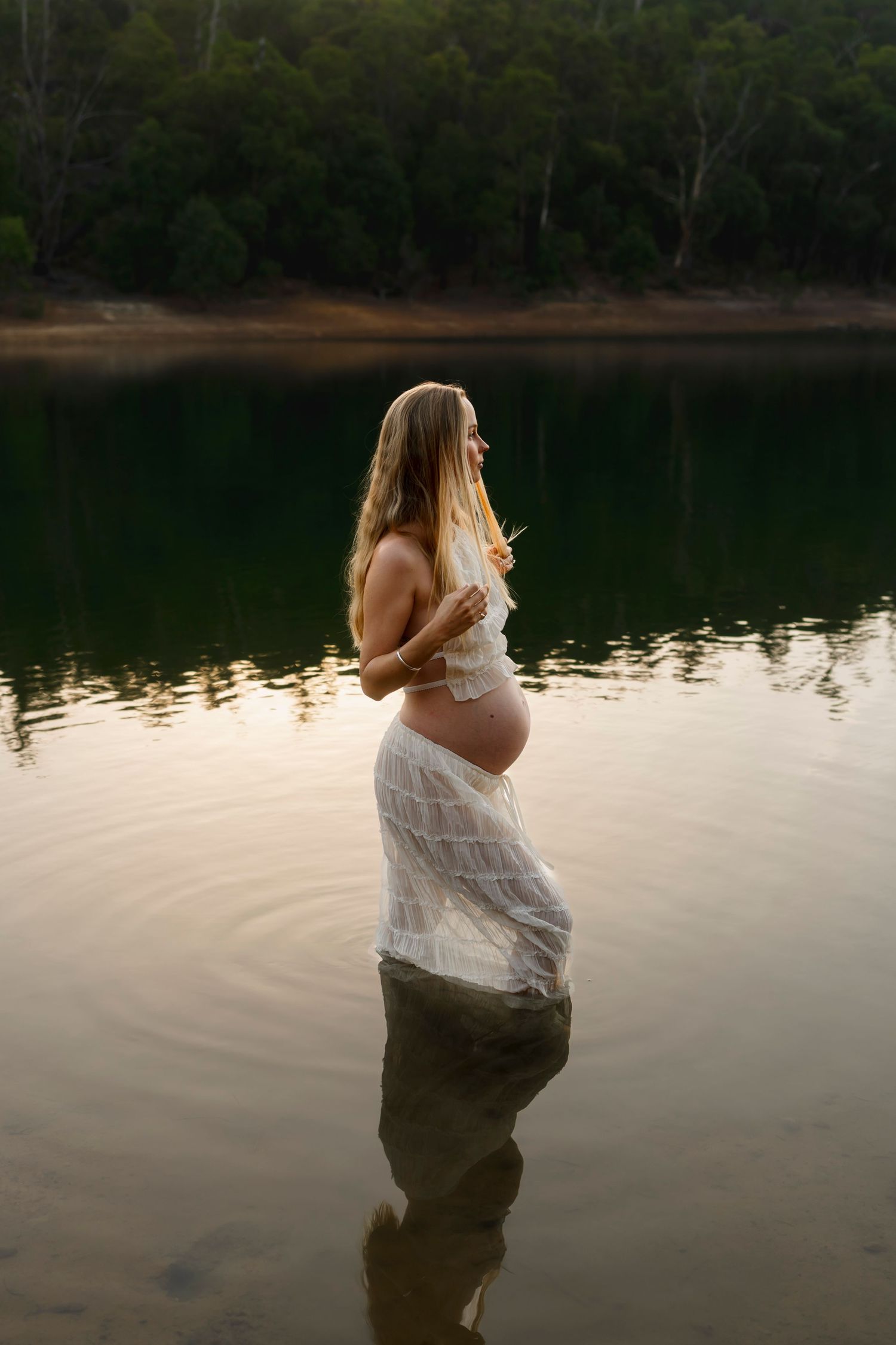 A person in a white dress stands in still water at sunset for a tranquil maternity photo session.